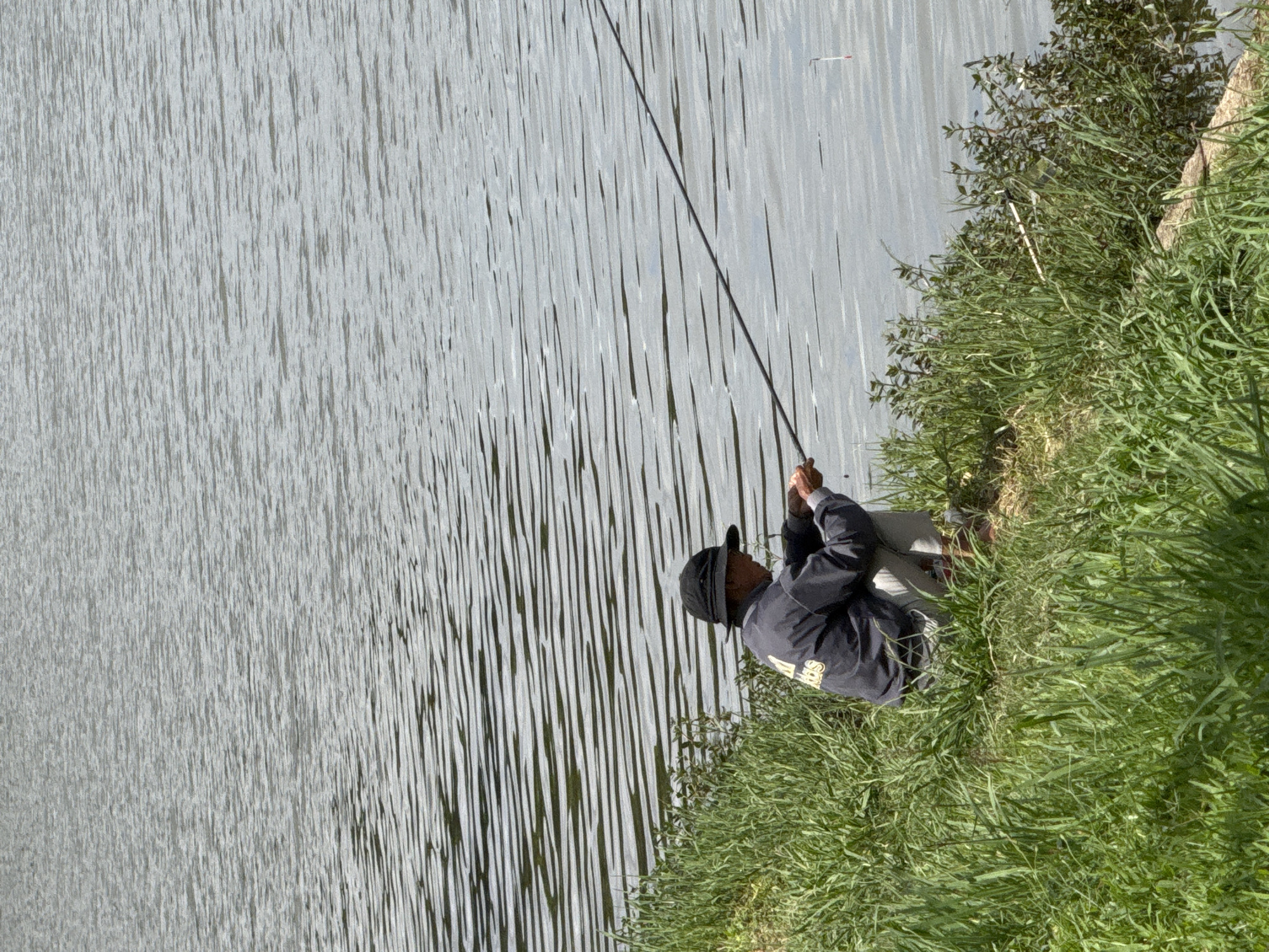A person wearing a black hat and dark jacket is fishing by a lake in Da Lat, Vietnam. The person is crouched on the grassy bank, holding a fishing rod. The water is calm with ripples, and the sky is overcast. Green grass and some small plants are visible along the shoreline.