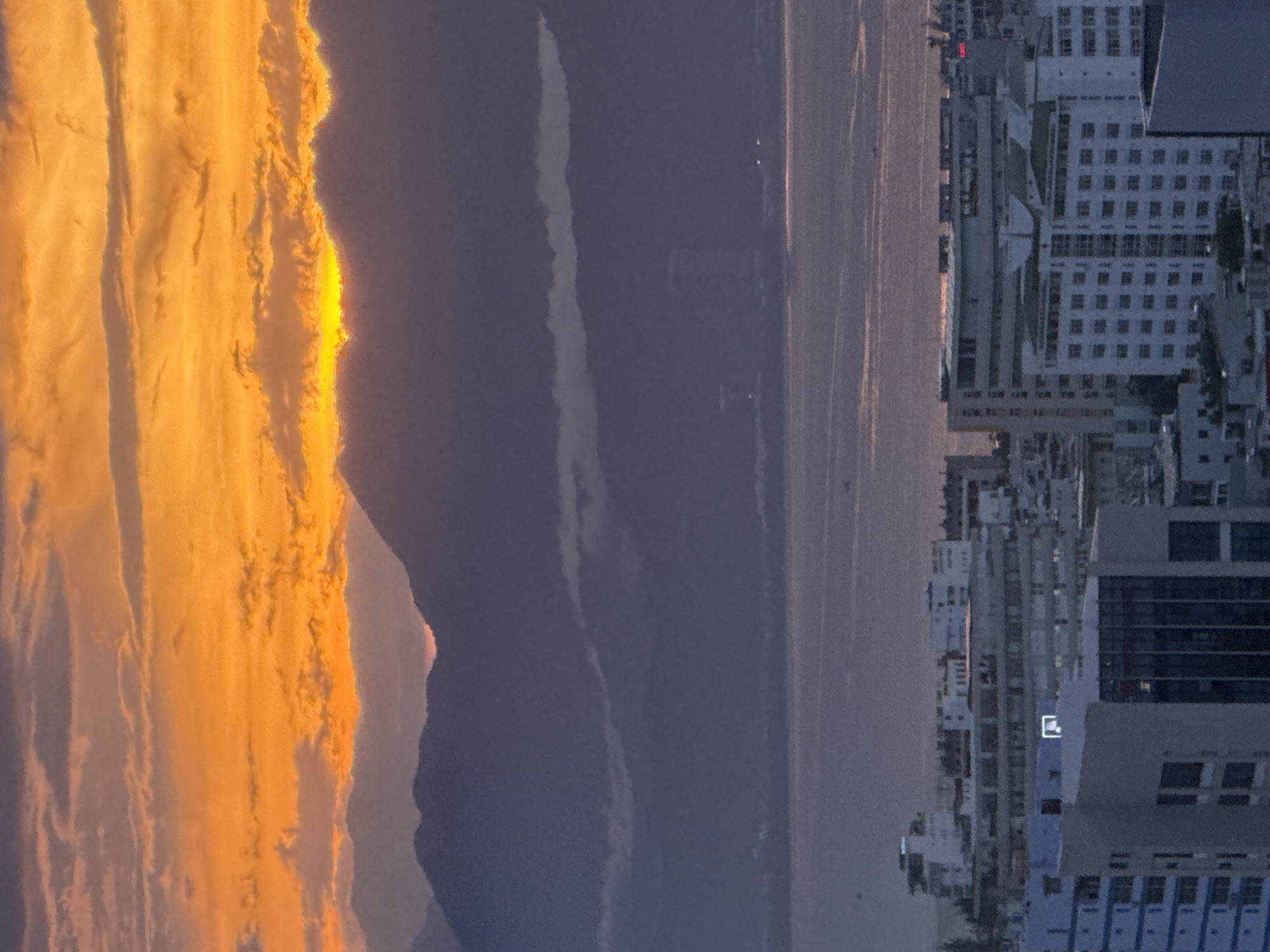 The image depicts a city skyline in Đà Nẵng, Vietnam, at dusk. The sky is ablaze with vibrant orange and yellow hues from the setting sun, casting a warm glow over the scene. Silhouetted against the dramatic sky are layers of mountains, their peaks softened by distance and atmospheric haze. In the foreground, the modern architecture of Đà Nẵng is visible, with numerous buildings of varying heights and designs. The presence of a body of water, likely the East Sea, is suggested by the expanse of calm water reflecting the sky's colors. The urban landscape comprises predominantly light-colored buildings, some with intricate window patterns, suggesting a mix of residential and commercial structures. A few smaller boats are faintly visible on the water. The overall impression is one of a tranquil yet dynamic urban setting at the transition from day to night.