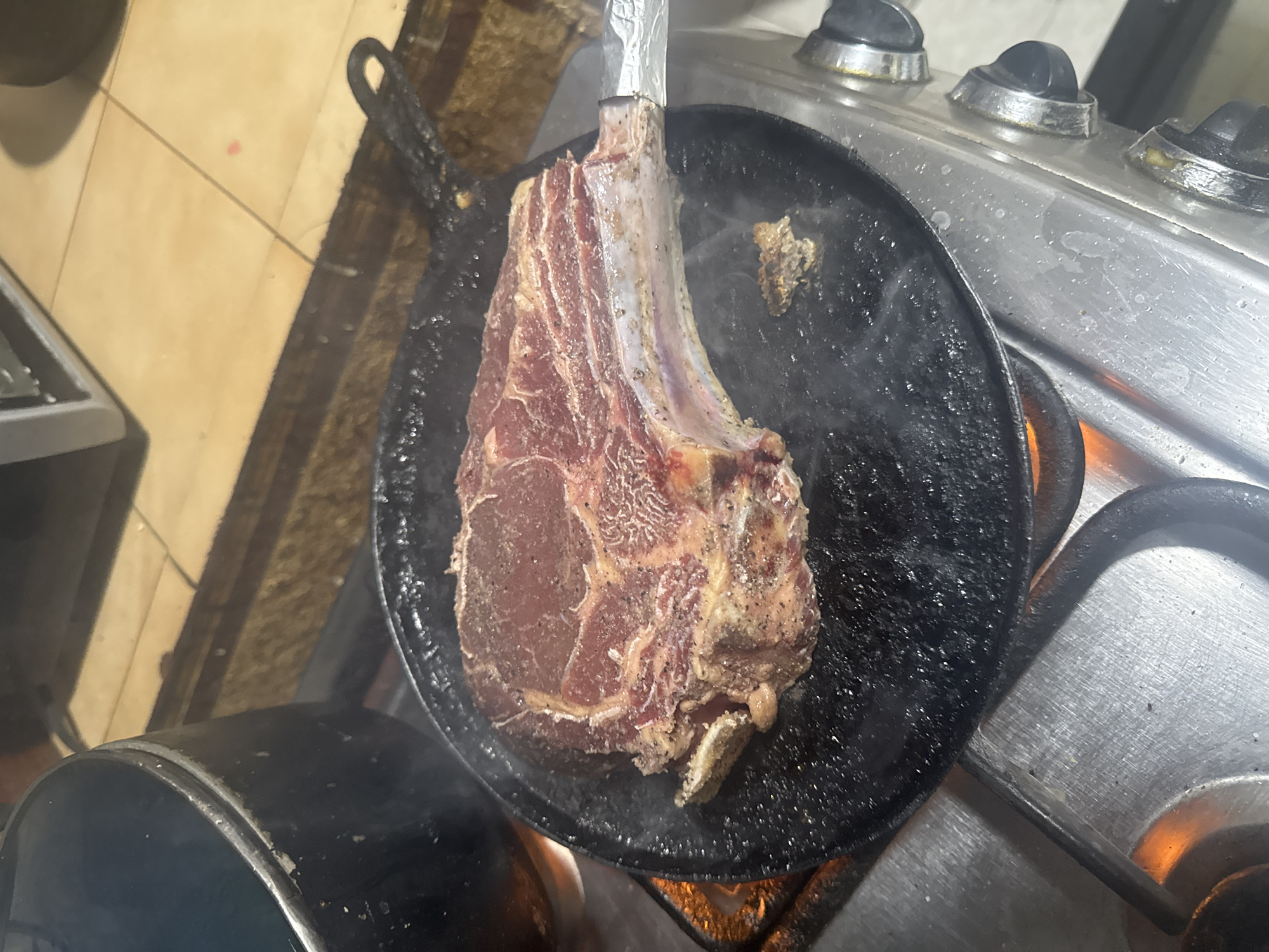 A raw, seasoned tomahawk steak rests in a black cast iron skillet atop a gas stove. The skillet is emitting steam. The stove is stainless steel with black knobs. The background shows tiled flooring and a metallic object. The location is possibly Maracay, Venezuela.
