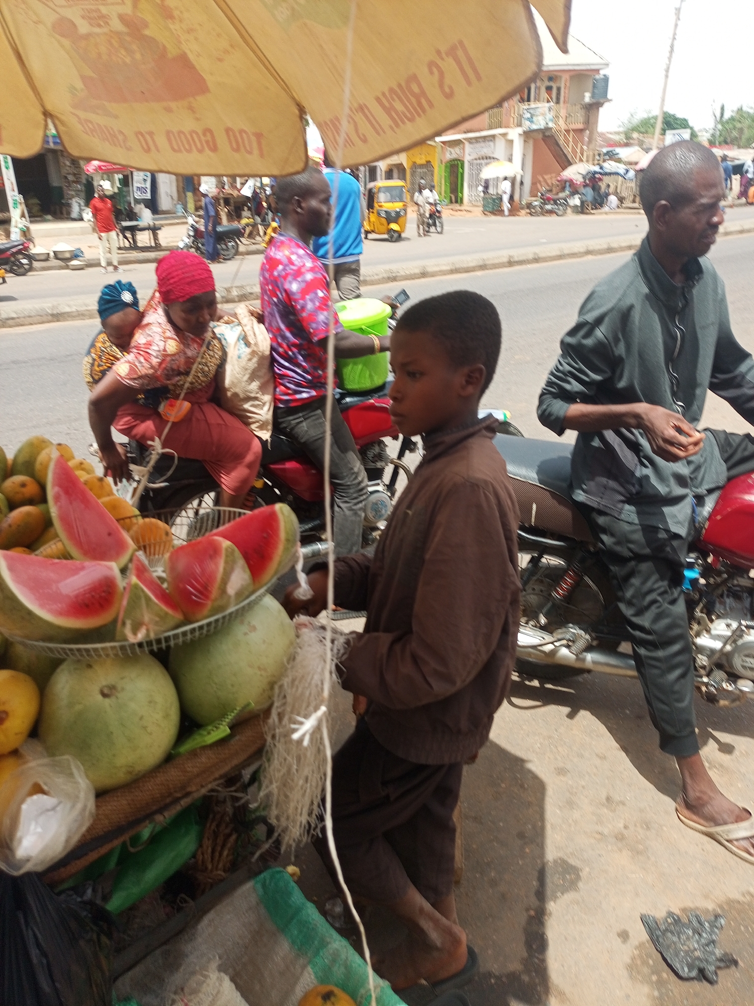 In Tempure, Nigeria, a street scene unfolds. A boy in a brown jacket stands near a fruit stand laden with mangoes, watermelons, and green gourds, some arranged on a metal rack. The stand is shaded by a yellow umbrella with advertising text. Nearby, a woman wearing a red headscarf and a baby on her back are seated on a red motorcycle. A man in a red patterned shirt stands beside them. Another man in a grey shirt and black pants is on a red motorcycle. In the background, traffic flows along the road, including motorcycles, auto rickshaws, and other vehicles. Buildings line the street.