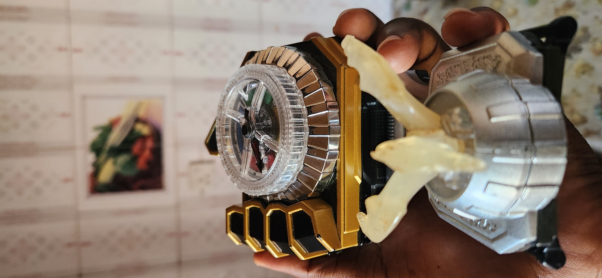 A close-up, slightly tilted shot captures a hand holding a complex, toy-like object. The object has a predominantly gold and black base, adorned with metallic, segmented detailing around a clear, circular component that resembles a spinning wheel. This wheel is divided into colored sections, including red and green, with a black pointer visible within. Extending from the object is a pale, translucent, wing-like protrusion with a vaguely organic shape, stained with a few small, orange marks. The background is out of focus, showing a patterned wallpaper or surface with a framed image of what appears to be a bouquet of flowers. The lighting suggests an indoor setting, possibly daytime given the natural light implied. The focus is entirely on the object and the hand, suggesting it is the primary subject of interest. No discernible text is visible on the object or in the background. The scene is neutral in terms of emotion.