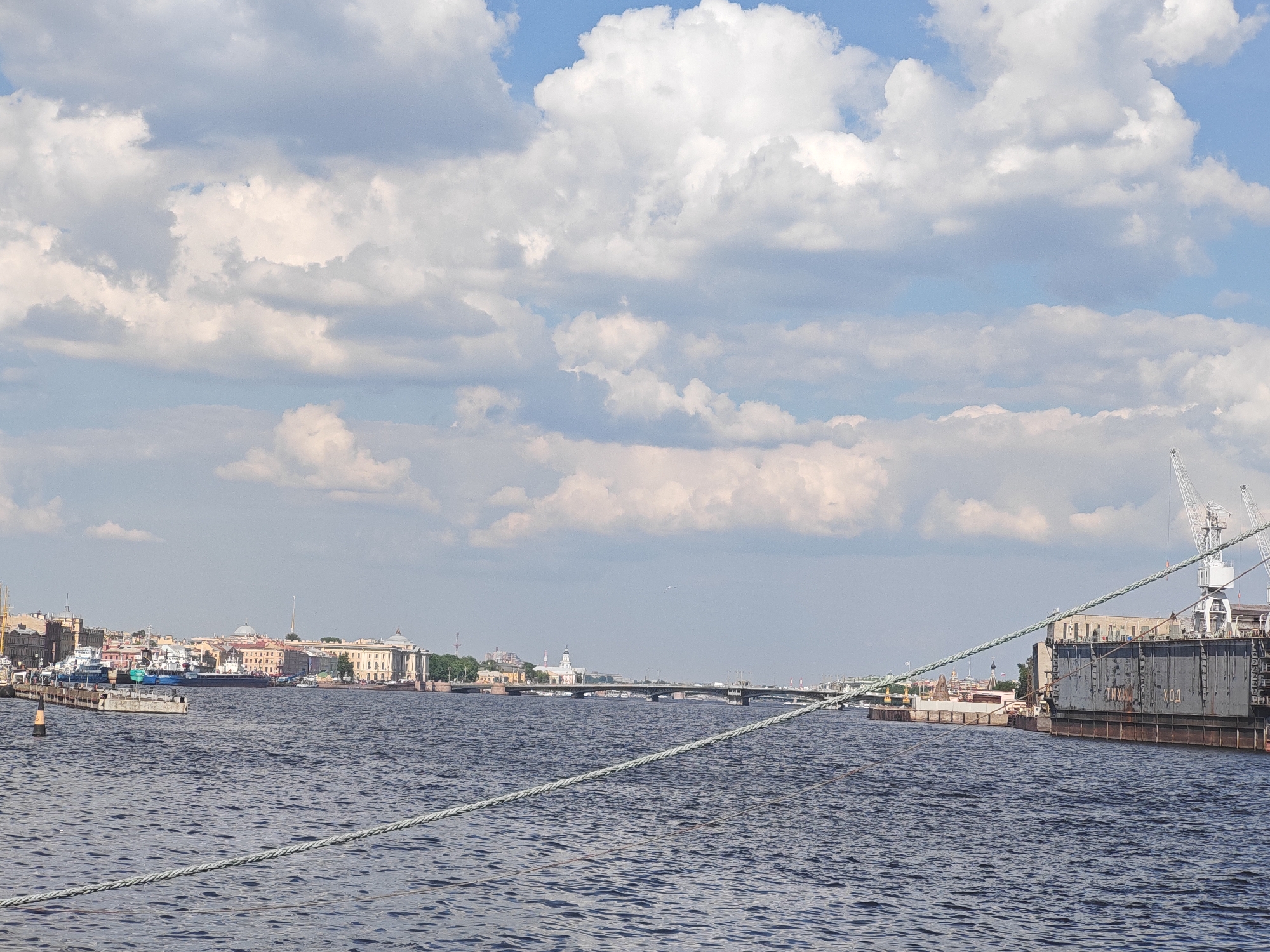 This wide-angle photograph captures a dynamic view of a river, likely the Neva, flowing through Saint Petersburg, Russia, under a bright, partly cloudy sky. The scene depicts a blend of historical urban architecture and active industrial maritime facilities.
In the foreground, the dark blue-grey river water is textured with numerous ripples, suggesting a gentle current. A thick, light-colored rope runs diagonally across the lower portion of the frame, indicating the vantage point is from a boat or pier. A black and yellow buoy is visible in the water on the left.
The left bank showcases a picturesque city skyline. A continuous line of classical European-style buildings, many with light-colored facades, some featuring domes and intricate architectural details characteristic of Saint Petersburg, stretches along the riverfront. Several smaller boats are docked close to the embankment, and more distant structures like spires are visible further downriver.
In the middle distance, a multi-span bridge, likely a drawbridge given the city's context, crosses the river, connecting the two sides. Beyond it, the city extends further into the horizon.
The right bank presents an industrial landscape. A large, dark-colored structure, possibly a floating dry dock or a large vessel undergoing maintenance, is moored along the shore. Two prominent white gantry cranes are positioned behind and on this structure, indicating ongoing port or shipbuilding activity. Cyrillic text is visible on the side of the large dark structure, appearing to read "ЧМХ-4" and "ХОД" (which translates to "Course" or "Movement").
The sky is a mix of clear blue patches and numerous large, puffy white cumulus clouds, suggesting fair weather and ample sunlight. The overall impression is one of a bustling port city, where historical grandeur coexists with active maritime industry. No individuals are clearly visible.