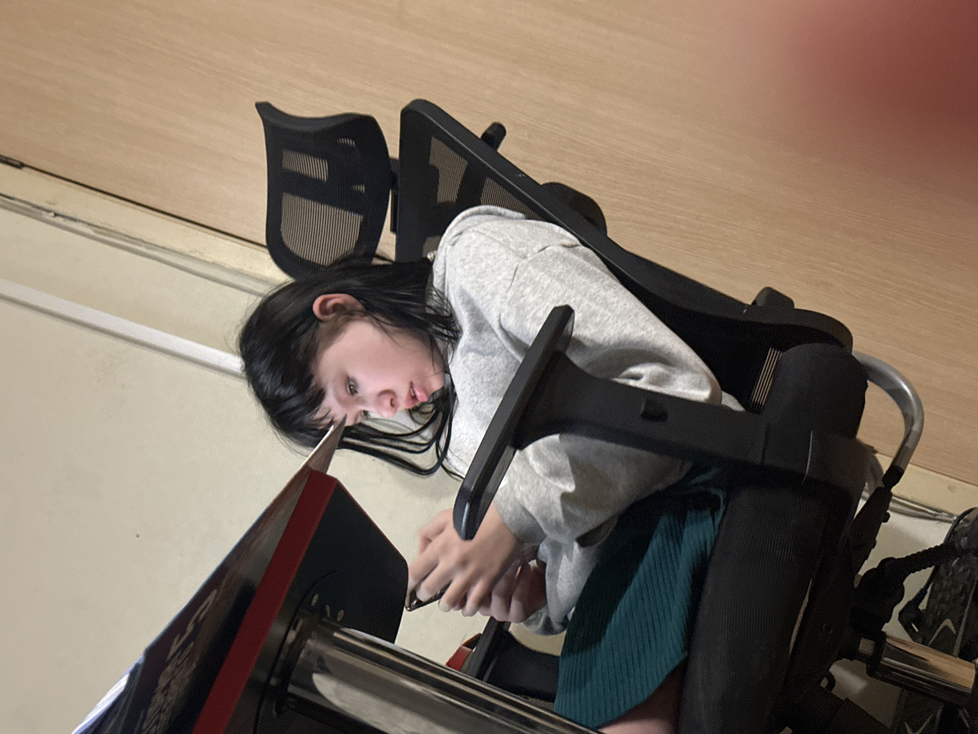 A young person is seated in a black office chair. They are wearing a grey sweatshirt and a dark green skirt. The individual is leaning forward and looking at an object on a surface in front of them. The background includes a wall and a light-colored floor. The chair has a mesh back and adjustable armrests. There is no evidence to indicate that the location is Moscow, Russia.