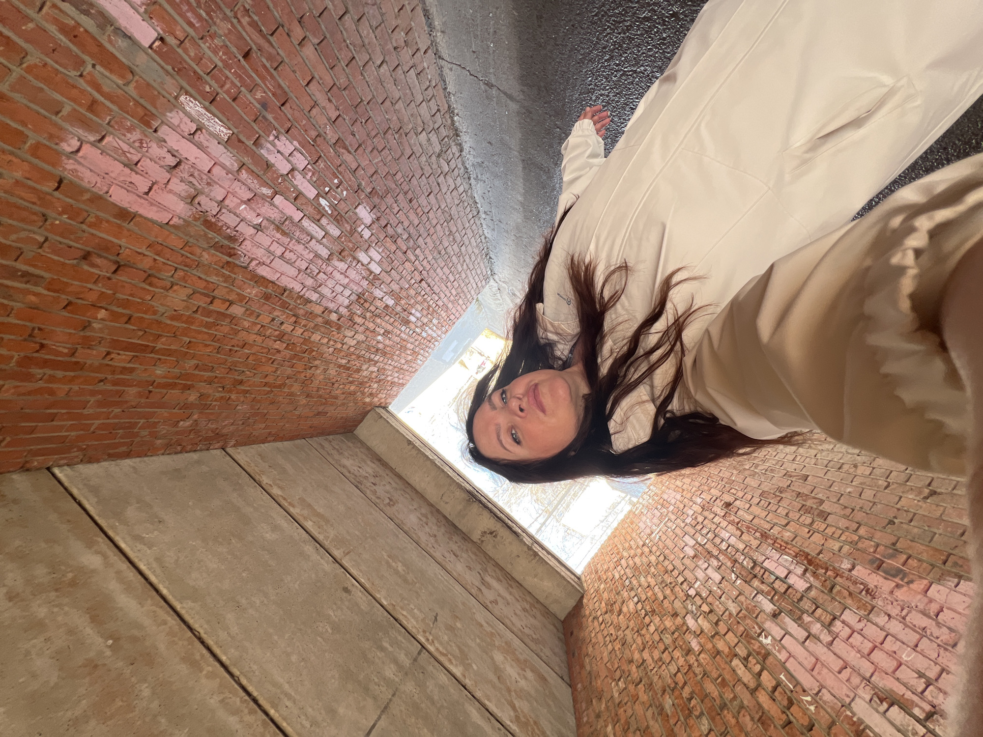 A woman with long, dark hair poses in what appears to be a concrete and brick underpass or tunnel. The walls of the tunnel are made of red brick, while the floor and ceiling seem to be constructed of concrete slabs. The woman is wearing a light-colored jacket or top. There is an opening at the far end of the tunnel, letting in natural light. The location of the image is unknown, and there is no evidence to suggest it is Noviy, Russia.