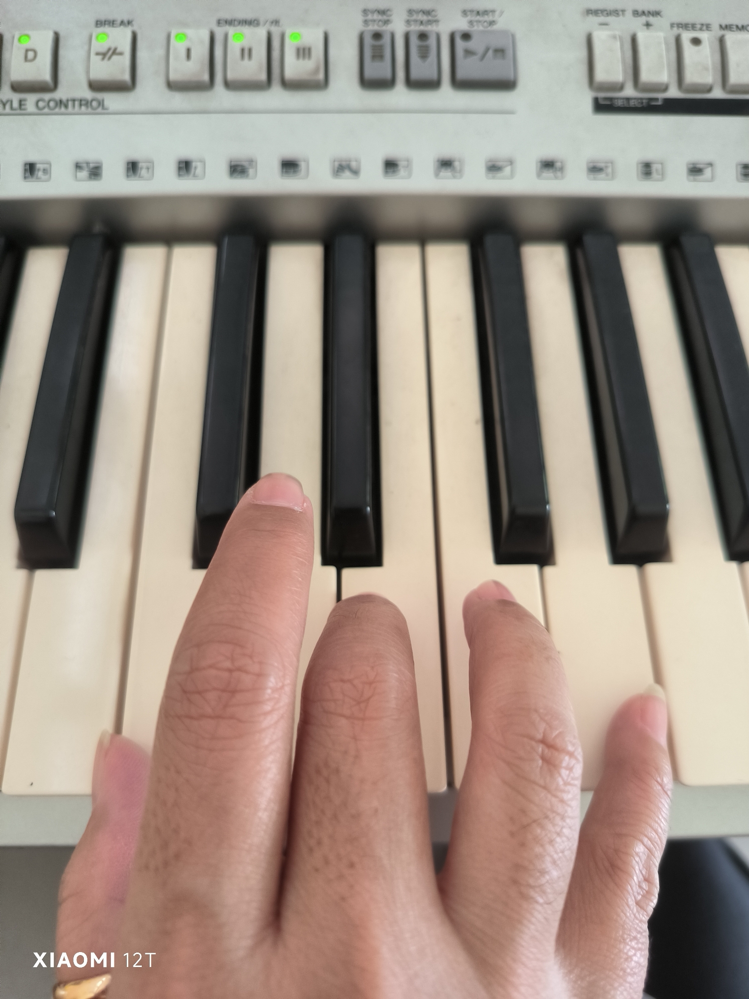 A person's hands are shown playing a keyboard. The keyboard has white and black keys. The person's left index finger is on a white key, and their thumb is resting on the black keys to the left of the white keys. Their right hand is partially visible, with fingers resting on white keys. Above the keys, there are various buttons and labels, including "BREAK", "ENDING", "SYNC STOP", "SYNC START", and "START/STOP". The image is taken from a top-down perspective, and the background is blurred. The bottom left corner of the image displays "XIAOMI 12T".