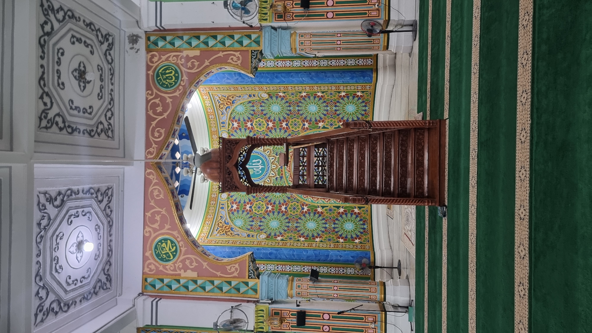 The image shows the interior of a mosque, likely in Keudekruenggeukueh, Indonesia, featuring a wooden minbar (pulpit) at the center, raised on a set of stairs. The minbar is ornately carved and topped with a dome. Behind the minbar, a colorful, geometrically patterned wall serves as a backdrop, adorned with Islamic calligraphy. The ceiling is decorated with intricate patterns. Green carpet covers the floor, marked with rows indicating prayer spaces. In the foreground, the carpet has decorative borders. Two ceiling fans are visible, one in the center and another to the right. There are also standing fans placed on either side of the room, and on the left wall, a microphone stand is present. The overall atmosphere suggests a well-maintained and decorated place of worship.