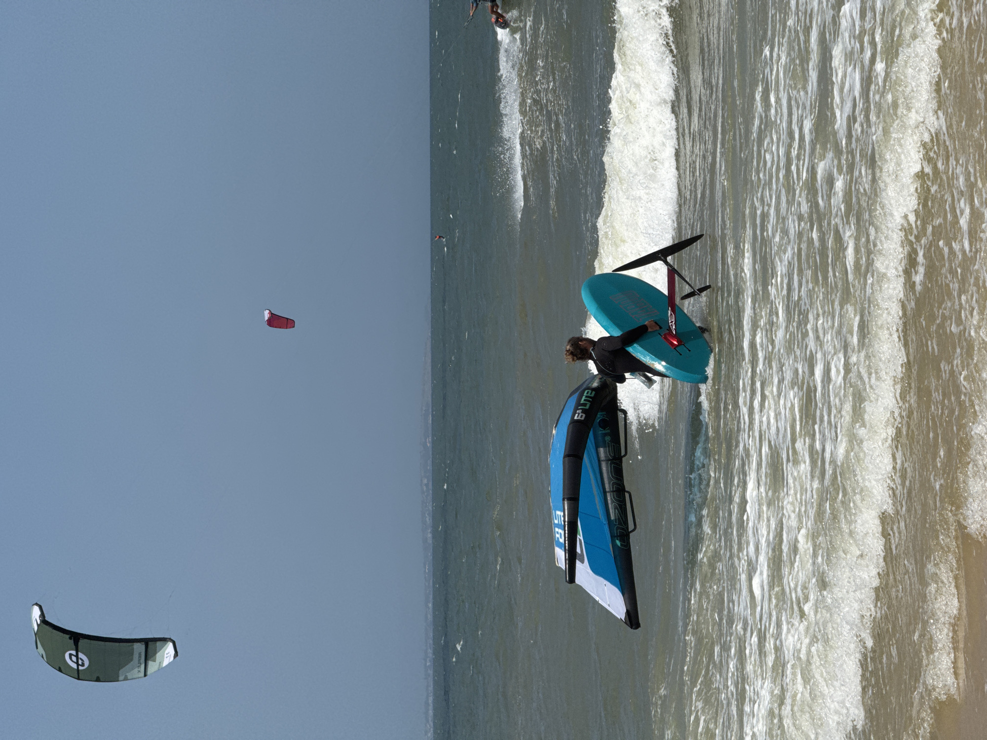 In Mũi Né, Vietnam, on a bright, hazy day, a person in a black wetsuit walks through the choppy, sunlit ocean, carrying a bright blue hydrofoil board and a large, blue and white wing. The wing, an Ozone brand kite, lies partially submerged in the water. The person is facing away from the camera, heading towards the shore. In the distance, other kitesurfers are visible on the water, with two more kites high in the sky. The ocean is a mix of dark and light blue, with white foam from waves crashing on the shore. The sky is a pale blue.