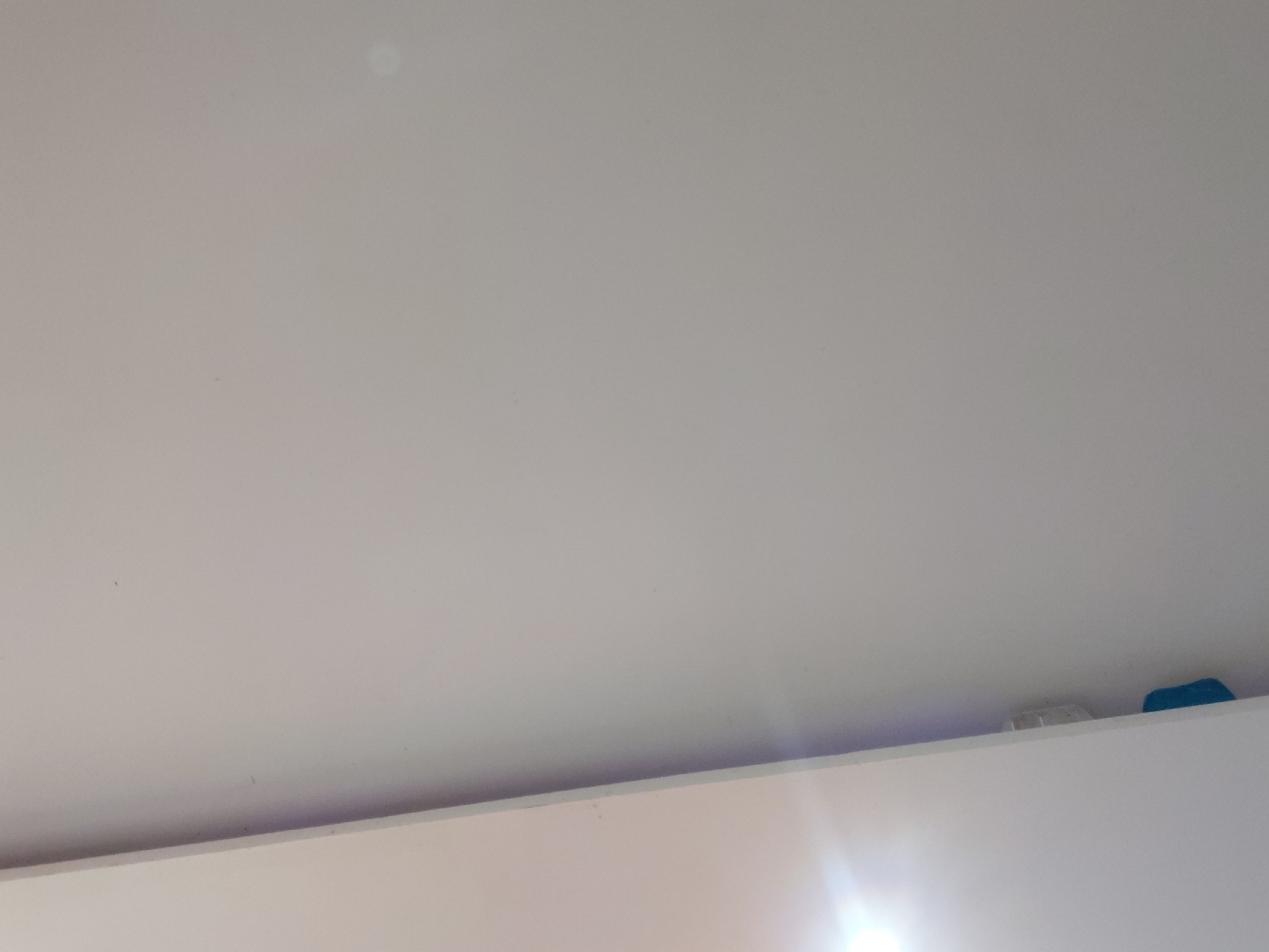 The image presents an upward-angled view of an indoor setting, dominated by a plain white or very light gray ceiling. The ceiling appears mostly smooth, with a few faint specks or minor imperfections visible across its surface. A crisp, horizontal line separates the ceiling from the top edge of a perpendicular wall, which occupies the bottom portion of the frame. This wall is also white or light-colored.
In the bottom right corner, resting on the upper edge of the wall, are two small objects. One is a white, somewhat rectangular object, possibly part of an electrical fixture or a small box. Adjacent to it is a bright blue object, which appears soft and amorphous, resembling a piece of fabric or a sponge. A significant lens flare or glare from an artificial light source is prominent in the bottom right, suggesting strong illumination originating from that area.
The scene is static, showing no human presence or discernible activity. The lighting implies an indoor environment, but the specific time of day is not evident. No visible text is present in the image. The setting depicts a simple interior space within Santa Ana, Venezuela.