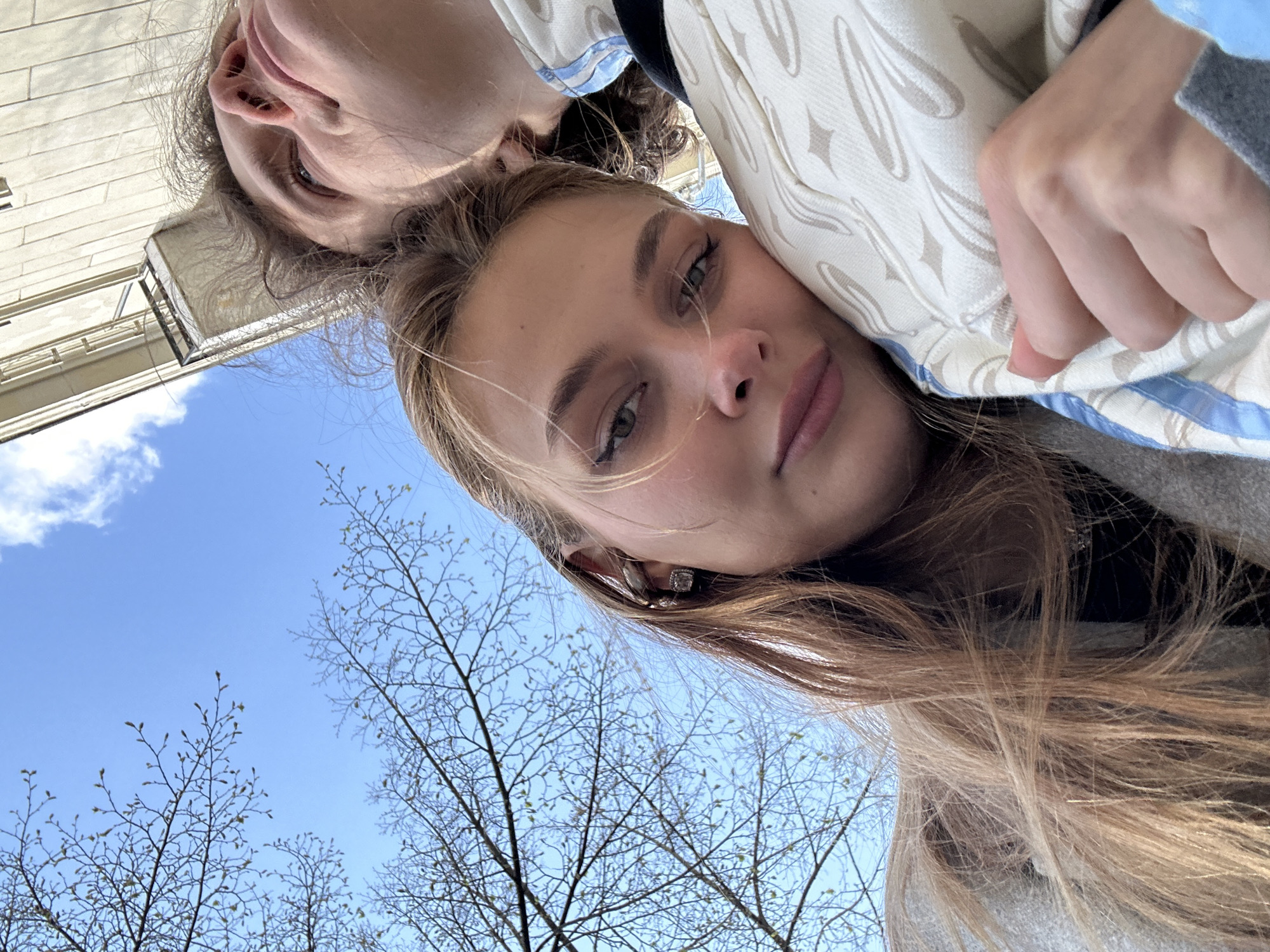 The image is a low-angle selfie of two people, likely taken outdoors. One person with long blonde hair is in the foreground, and another person is positioned slightly behind and above. The background features a clear blue sky and bare tree branches with small buds, suggesting early spring. The building details indicate an urban environment. Given the location of Krasnoyarsk, Russia, it is plausible the photo was taken within a park or urban area in this city.