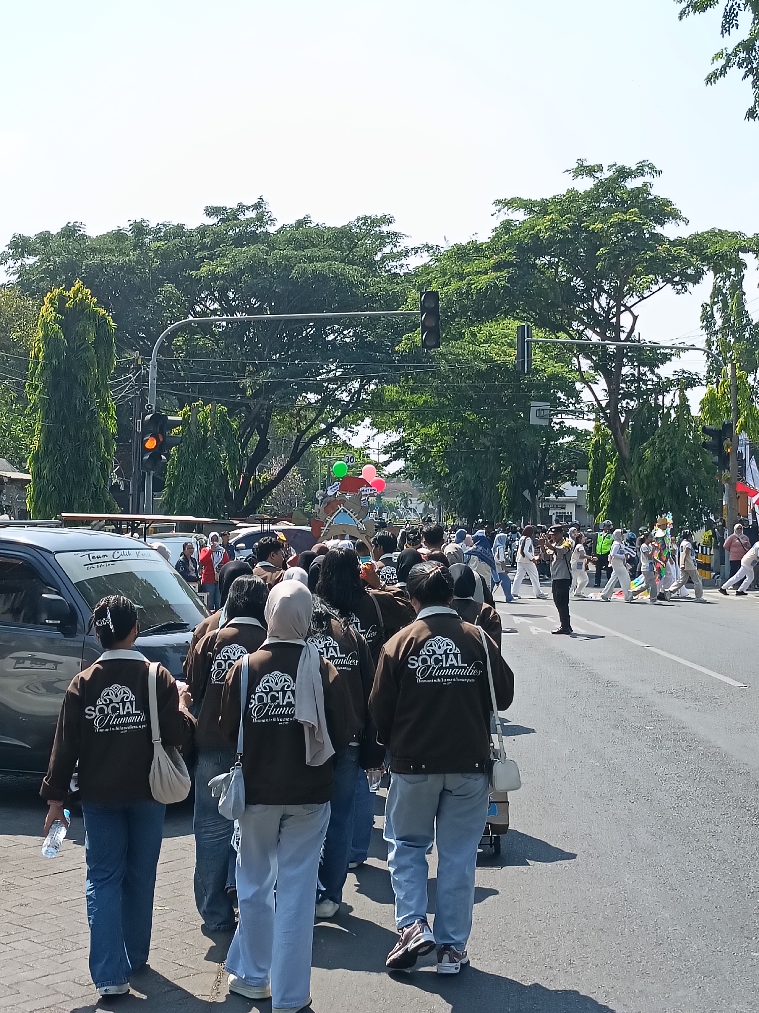 The image depicts a sunny street scene in Kediri City, Indonesia, where a group of people, likely students, are walking in formation along the road. They are wearing matching brown jackets with a white logo and text that appears to say "SOCIAL Humanities" and "Humanism adalah anugerah". Some individuals are carrying bags, and one person is holding a water bottle. In the background, there are trees, traffic lights, a vehicle, and other pedestrians. The overall atmosphere suggests a public event or parade.