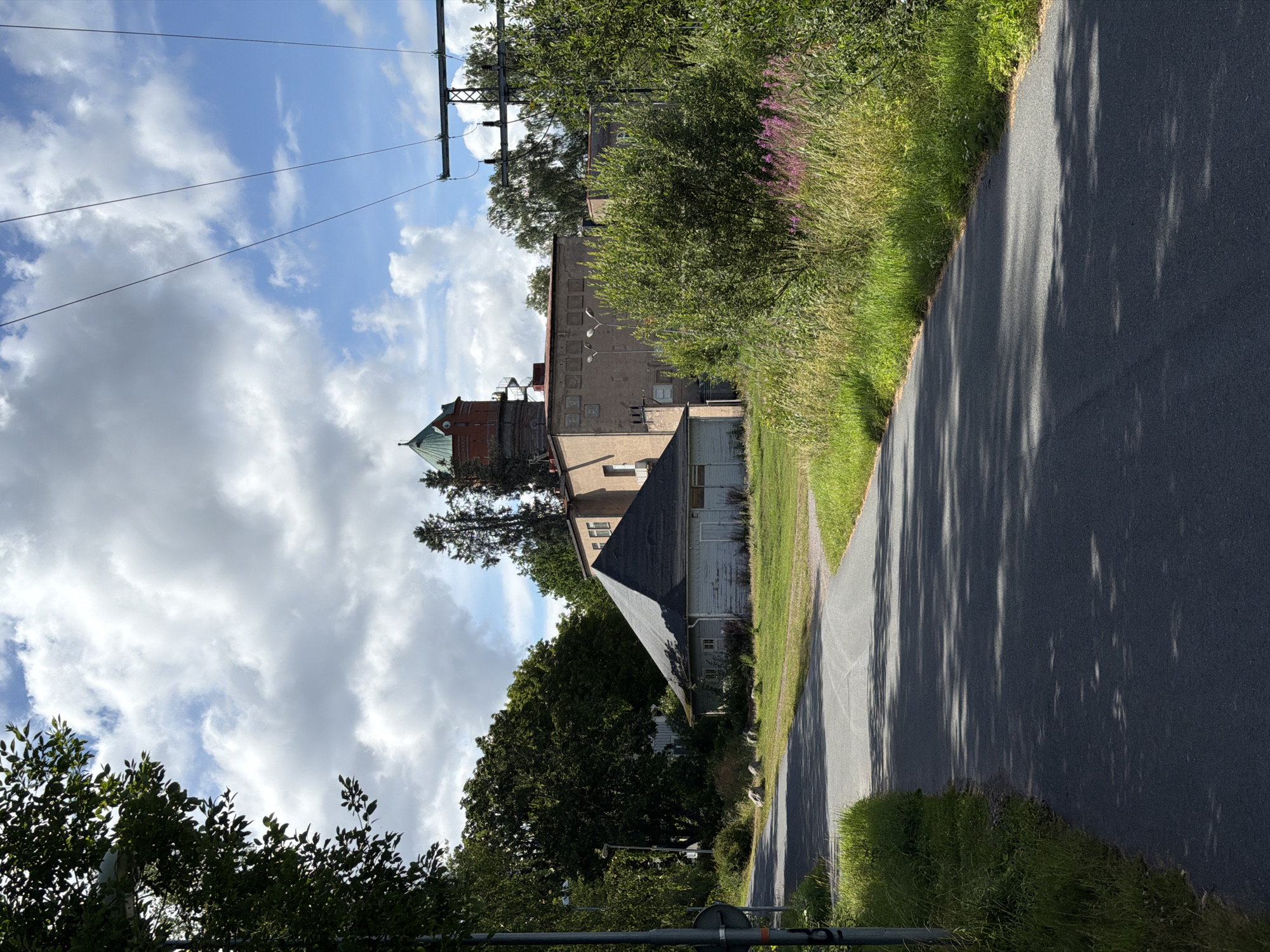 A rural paved road curves to the right through a grassy area with trees. On the left, a dark gray pole with a round sign is partially visible. In the background, a tan building with a red-brick tower topped by a green roof stands to the right of a smaller, dark-colored building with a slanted roof. Tall green trees and bushes surround the structures, and power lines are visible against a blue sky with white clouds. The scene is lit by daylight.