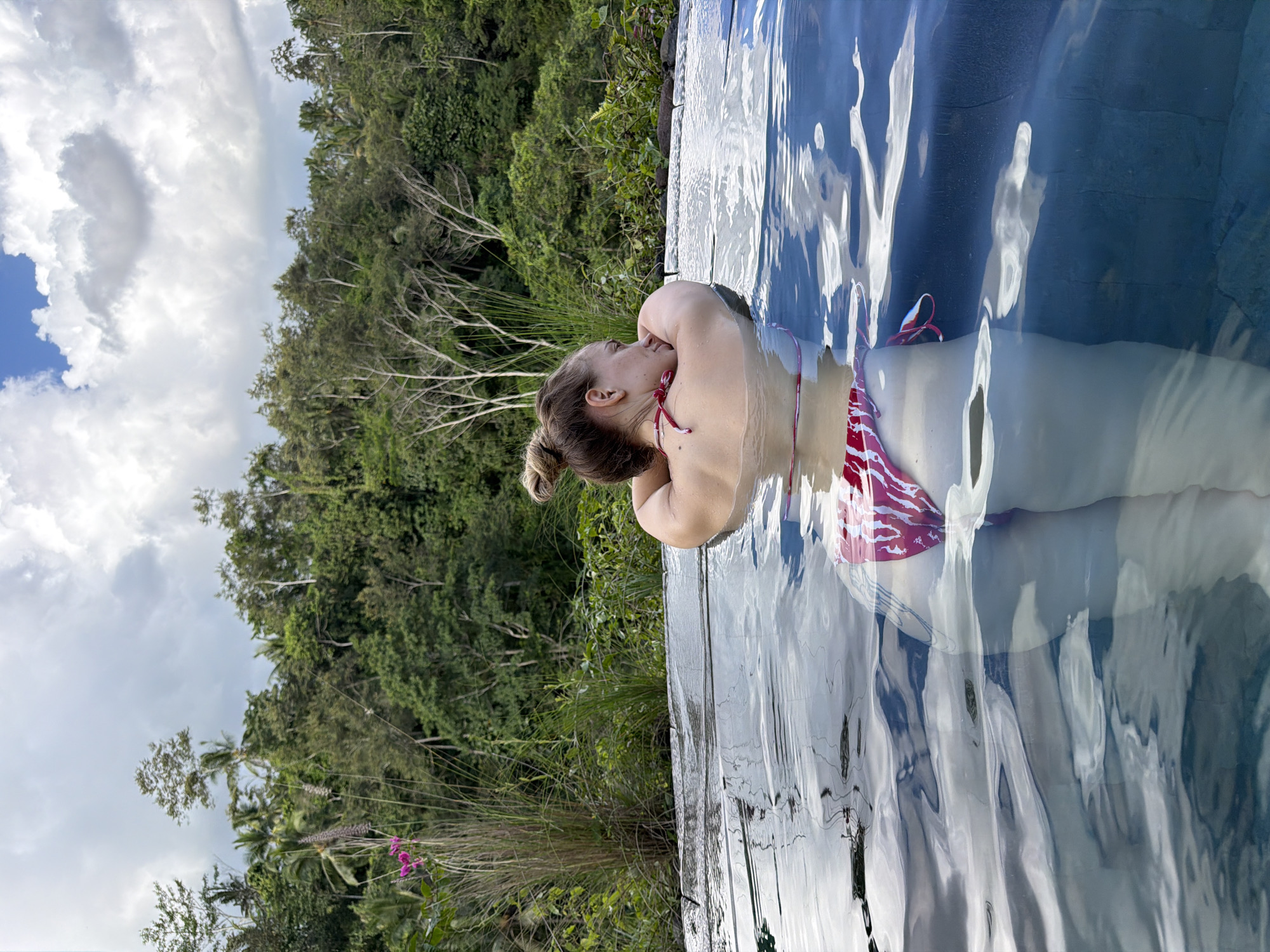 A woman with blonde hair in a bun, wearing a red and white patterned bikini, is in a swimming pool. She is facing away from the camera, resting her head on her arms, which are on the edge of the pool. The pool is made of dark material, possibly stone, and appears to be an infinity pool. Water ripples on the surface. The background is a lush, green jungle with tall trees and vegetation. The sky above is cloudy with some patches of blue. This scene is located in Tegallalang, Indonesia, known for its scenic rice terraces and tropical landscapes.