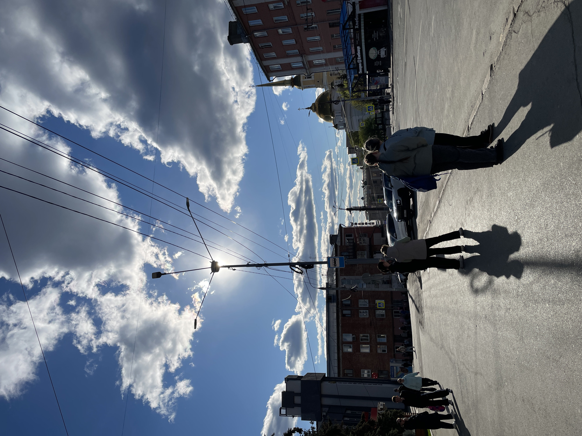 The image depicts a street scene in Izhevsk, Russia, notable for its clear sky dotted with cumulus clouds. An orthodox church with a golden dome and a tall spire is visible in the distance. Overhead electrical lines crisscross the sky. A street light is positioned mid-frame, with the sun shining brightly behind it. Pedestrians can be seen walking on the sidewalk and street, casting long shadows. Buildings of varying heights and architectural styles line the street, including brick and modern structures. A bus stop with signage in Russian is also present.
