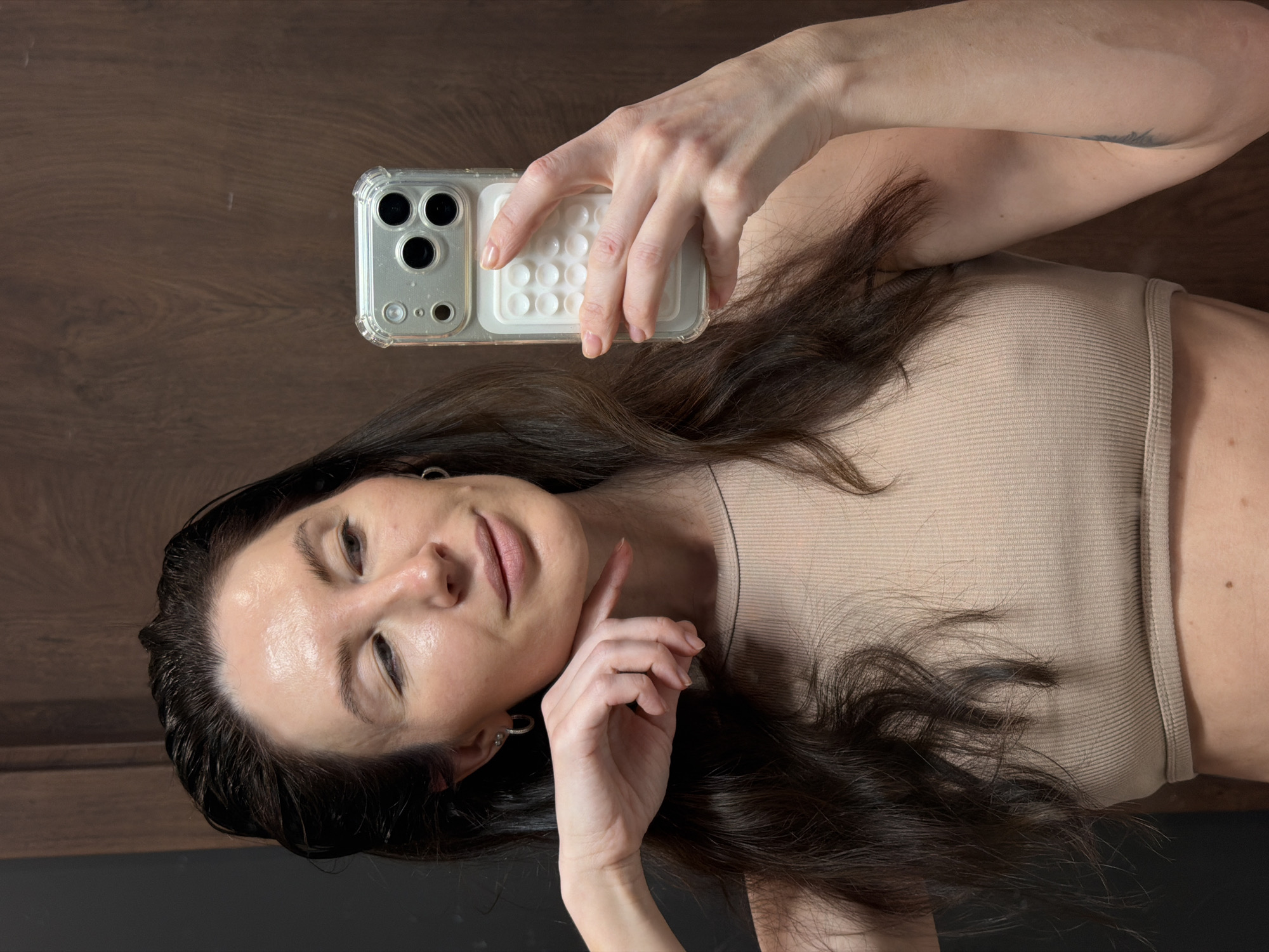 A woman with dark hair and fair skin takes a selfie using a smartphone with a clear case. The background is a wooden surface. She is wearing a beige top. There is no indication of the location being Perm, Russia.