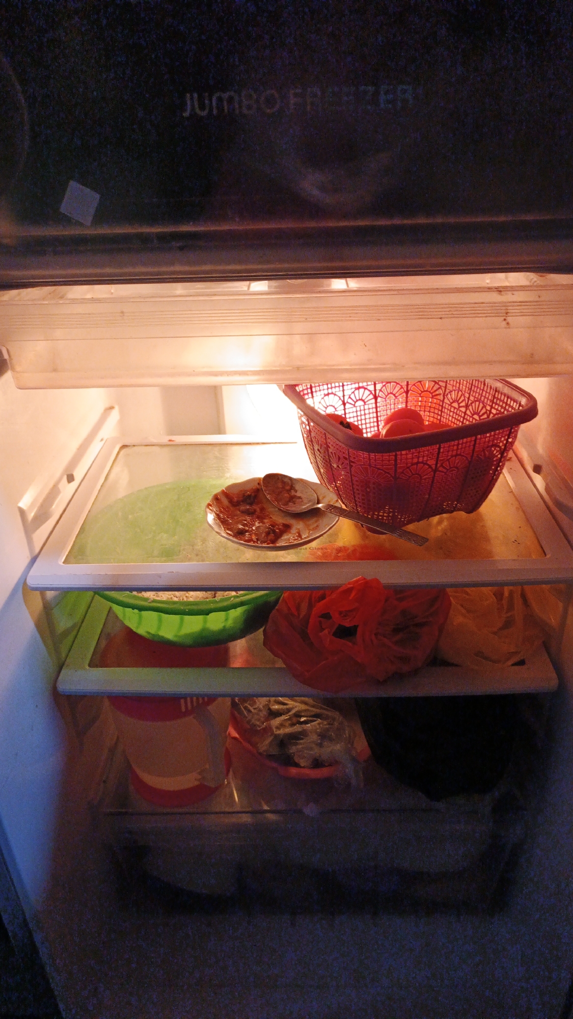 The image depicts the interior of a refrigerator. On the top shelf, a red plastic basket contains several red tomatoes. To the left of the basket is a small white plate with remnants of food and a spoon. Below this shelf, a green bowl holds what appears to be rice. To the right of the green bowl are two plastic bags, one red and one yellow. Further down, on the shelf below, are various containers, including a pink and white cylindrical container and a clear plastic bag containing food. The overall environment appears to be cluttered and somewhat unorganized. The text "JUMBO FREEZER" is visible at the top of the refrigerator. This refrigerator is located in Gunungreja, Indonesia.