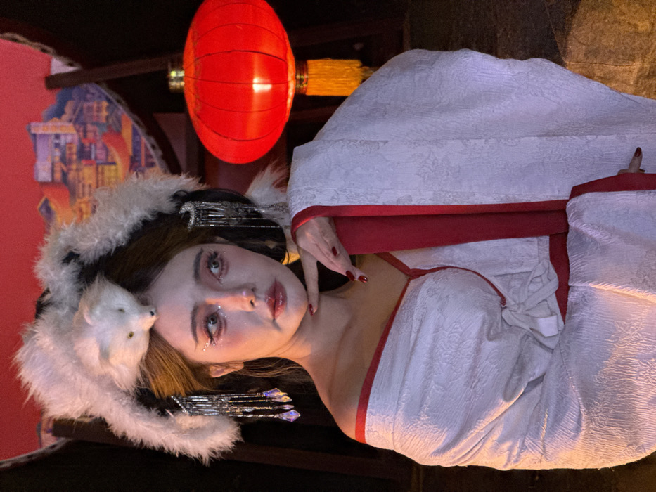 An individual, identified as female, is depicted in a medium close-up shot within a dimly lit environment, likely in Yuzhong District, China. The subject has dark hair styled with a prominent white faux-fur headpiece resembling an animal, and adorned with multiple silver-toned hair ornaments featuring crystal-like drops on both sides of the head. Detailed facial makeup is visible, including glitter on the eyelids and under eyes, winged eyeliner, rosy blush, and glossy lips. She is wearing a white robe or dress with a subtle patterned texture and prominent red trim along the neckline and front opening, styled off-shoulder to expose her right shoulder. A red sash or tie is also visible at her waist. Her right hand, with red painted fingernails, is positioned with fingers gently touching her chin and jawline. Her gaze is directed upwards and to the right of the frame. In the background, to the right of the subject, a large, illuminated red Chinese lantern with a gold tassel hangs. Behind the subject's head, a decorative panel or screen features an illuminated illustration of traditional Chinese architecture.
