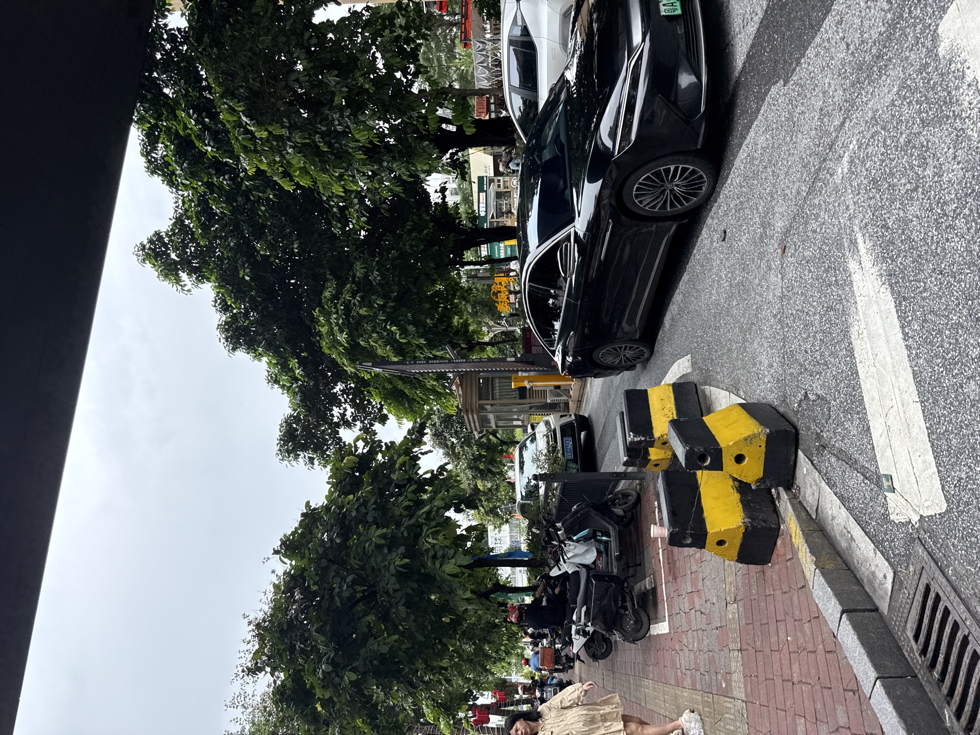 The image depicts a street scene in Haizhu District, China, with a focus on a black sedan parked on the right side of the road. The car is angled slightly away from the viewer, and its license plate, which appears to be green with white characters, is partially visible. In the foreground, yellow and black concrete barriers are placed on the roadside. The street is paved with asphalt, and white markings are visible on the road surface.
To the left of the road is a sidewalk made of red brick pavers. Several motorcycles and scooters are parked along the sidewalk. A woman in a light-colored dress is walking on the sidewalk, moving away from the viewer. In the background, lush green trees line the street, and a few buildings are visible. The sky is overcast. A dark, horizontal structure, possibly an overhang or bridge, is present at the top of the frame.