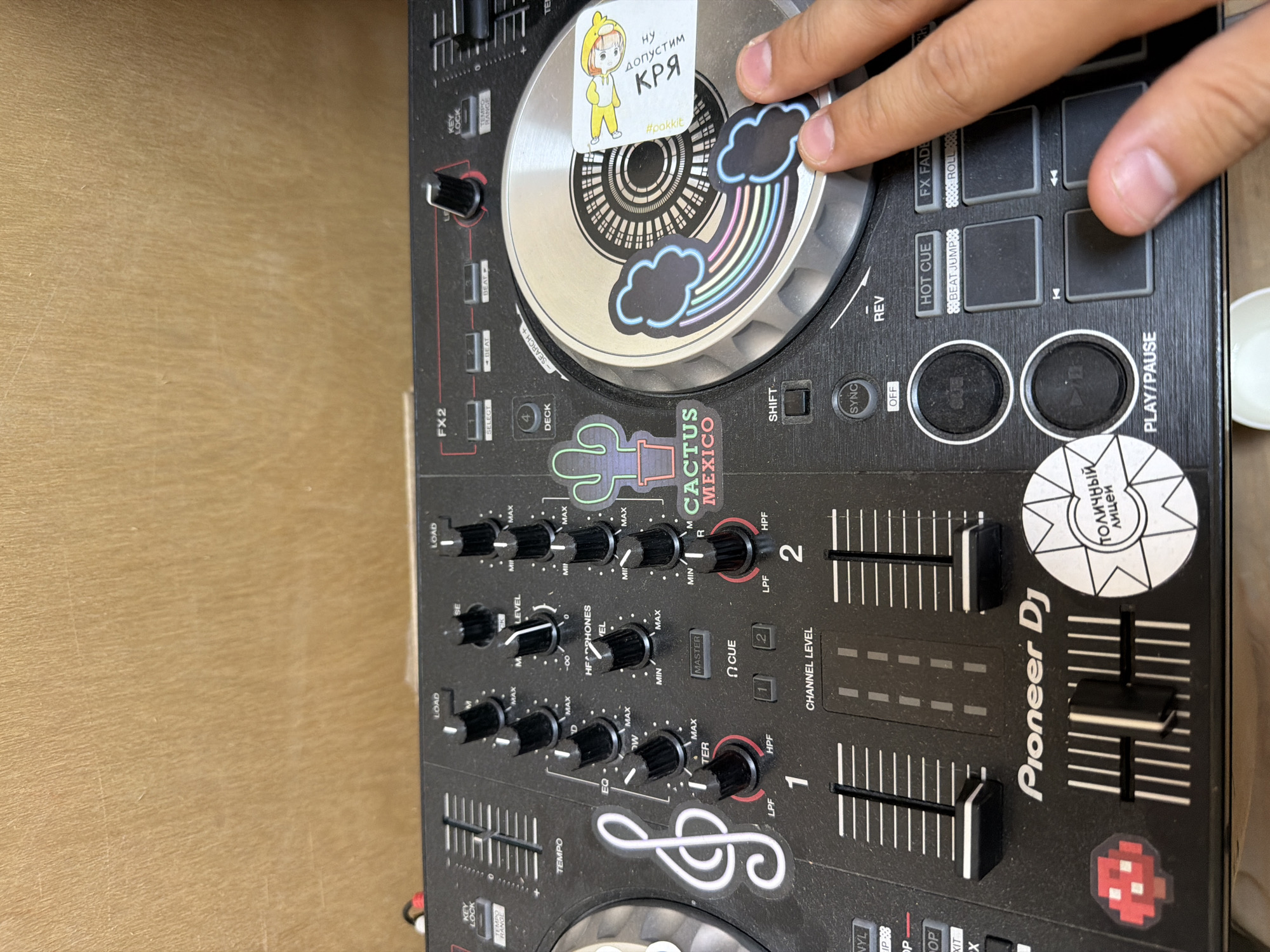 The image shows a close-up view of a Pioneer DJ controller, possibly a DDJ model, set on a wooden surface. A hand is touching the jog wheel, which is adorned with stickers: one of a cartoon character and another of dark clouds with a rainbow. The controller itself has several other stickers, including a cactus with "CACTUS MEXICO" written below, a bass clef, a red pixelated character, and a circular sticker with Cyrillic text "ТОЛИЧНЫЙ ЛИЦЕЙ". The controls visible include knobs, faders, buttons labeled with functions like "FX FADE", "HOT CUE", "PLAY/PAUSE," "SYNC," "SHIFT" and controls for EQ, headphone level, and channel levels. The presence of Cyrillic text suggests the controller is located in a Russian-speaking area, and the school name printed on the sticker might provide more specific geographical location.