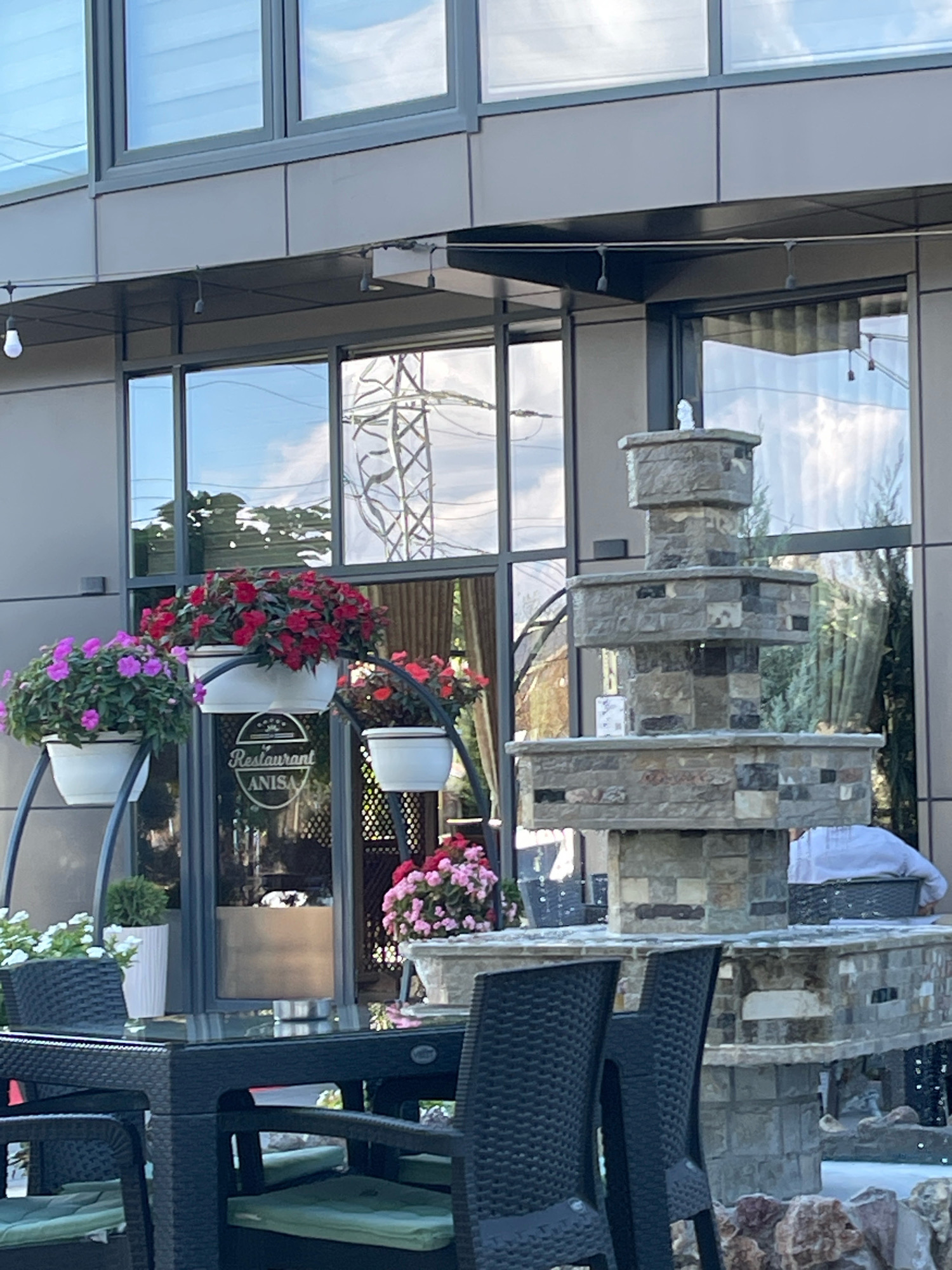 The image shows a restaurant called "Anisa" located in Mirushë, Kosovo. The exterior of the restaurant features large windows reflecting the sky and surrounding trees, along with a multi-tiered stone fountain. In front of the restaurant, there are tables and chairs set up for outdoor dining. Several potted plants with pink and red flowers are strategically placed around the seating area. A person wearing a light-colored garment is visible in the background, seated in one of the chairs.