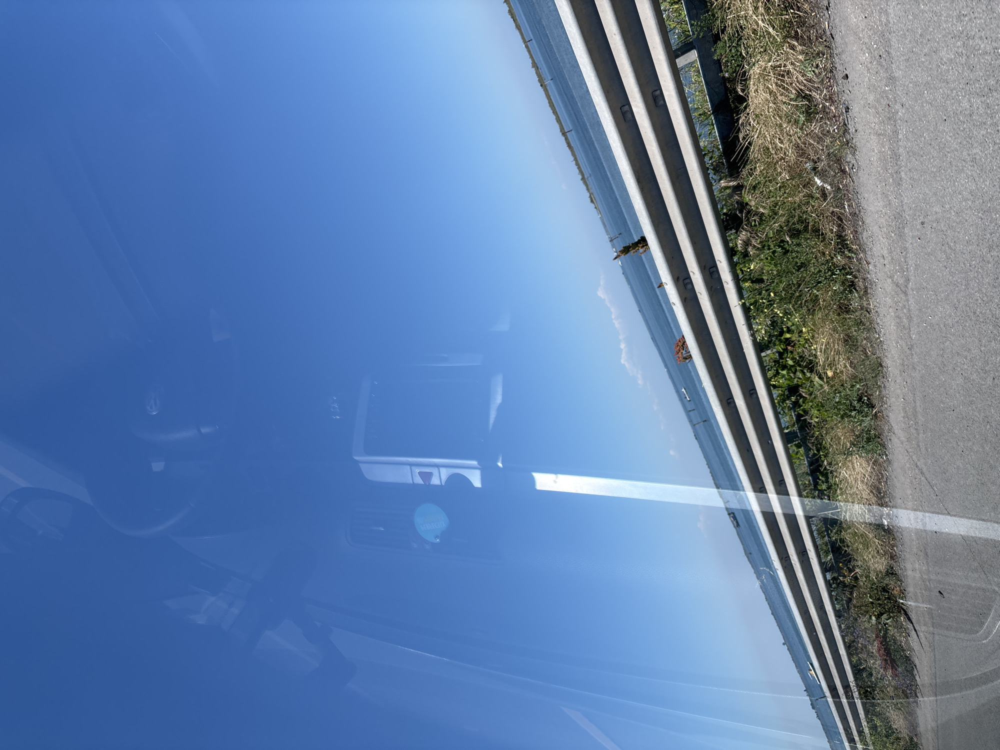 The image appears to be taken from inside a vehicle, with the windshield acting as a reflective surface. The reflection primarily shows a clear, blue sky. Outside the vehicle, visible through the windshield, is a section of road with a metal guardrail on the side. Vegetation grows along the edge of the road. Details inside the vehicle, such as parts of the dashboard and rearview mirror, are also visible in the reflection. Without more details, it is not possible to find this location in Chioggia, Italy.