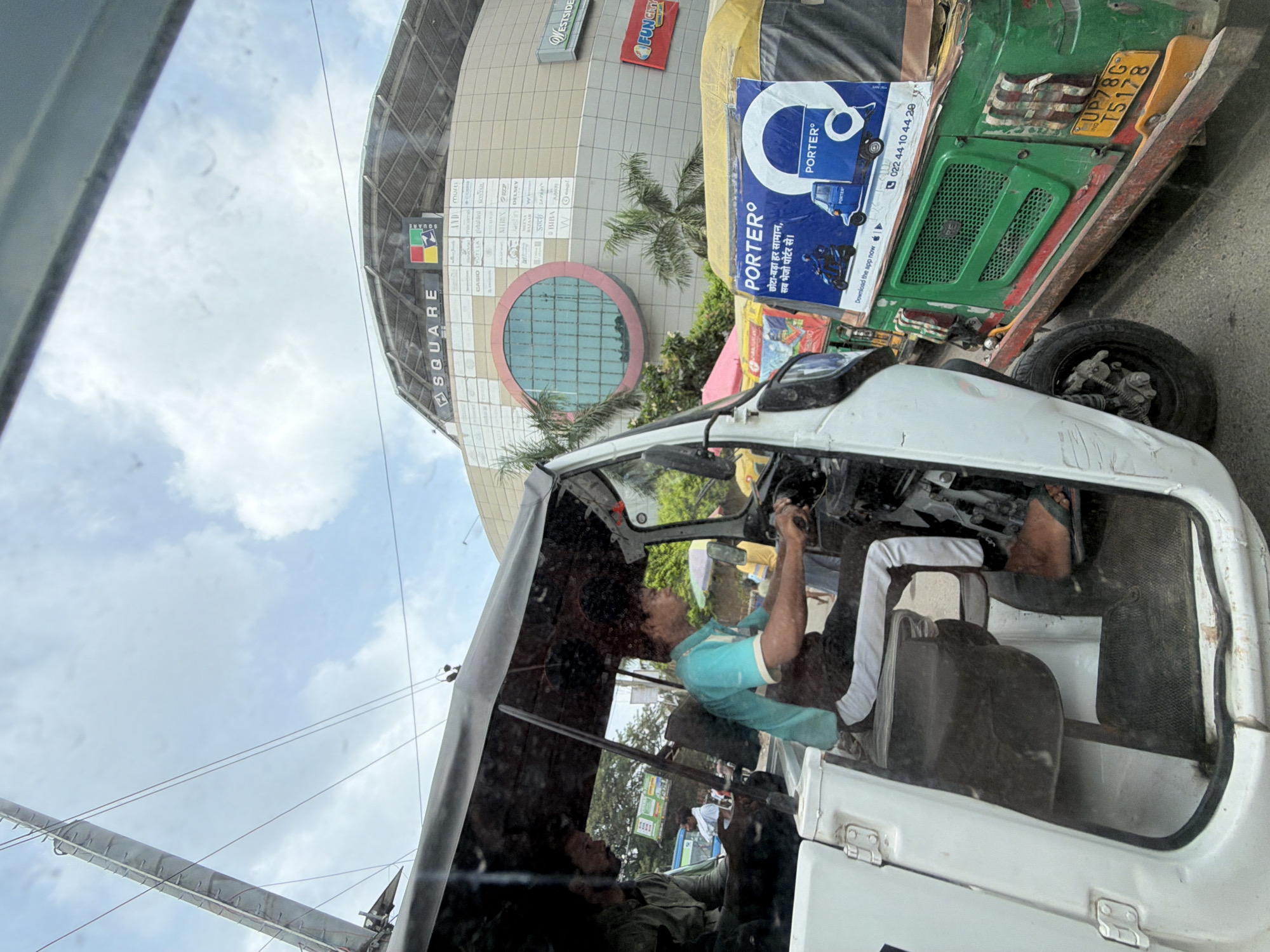 The image captures a street scene in Kanpur, India, with a focus on the interiors of two auto-rickshaws. In the foreground, a white auto-rickshaw is partially visible, showing its open door and interior. A man wearing a blue t-shirt and black pants with white stripes is seated in the driver's position, gripping the steering wheel. Another person, partially obscured, is visible in the passenger seat behind him. Outside the white auto-rickshaw, a green auto-rickshaw is parked alongside. This green auto-rickshaw displays a large advertisement for "PORTER°" with text in Hindi and English, featuring images of a scooter and a delivery truck. The license plate of the green auto-rickshaw is visible, showing "UP 78G T5178". In the background, a large building with a curved roof and circular window is visible, identified by a sign as "SQUARE". The sky above is cloudy. The overall scene suggests a busy urban environment, possibly a commercial area or a traffic intersection, with several auto-rickshaws present.