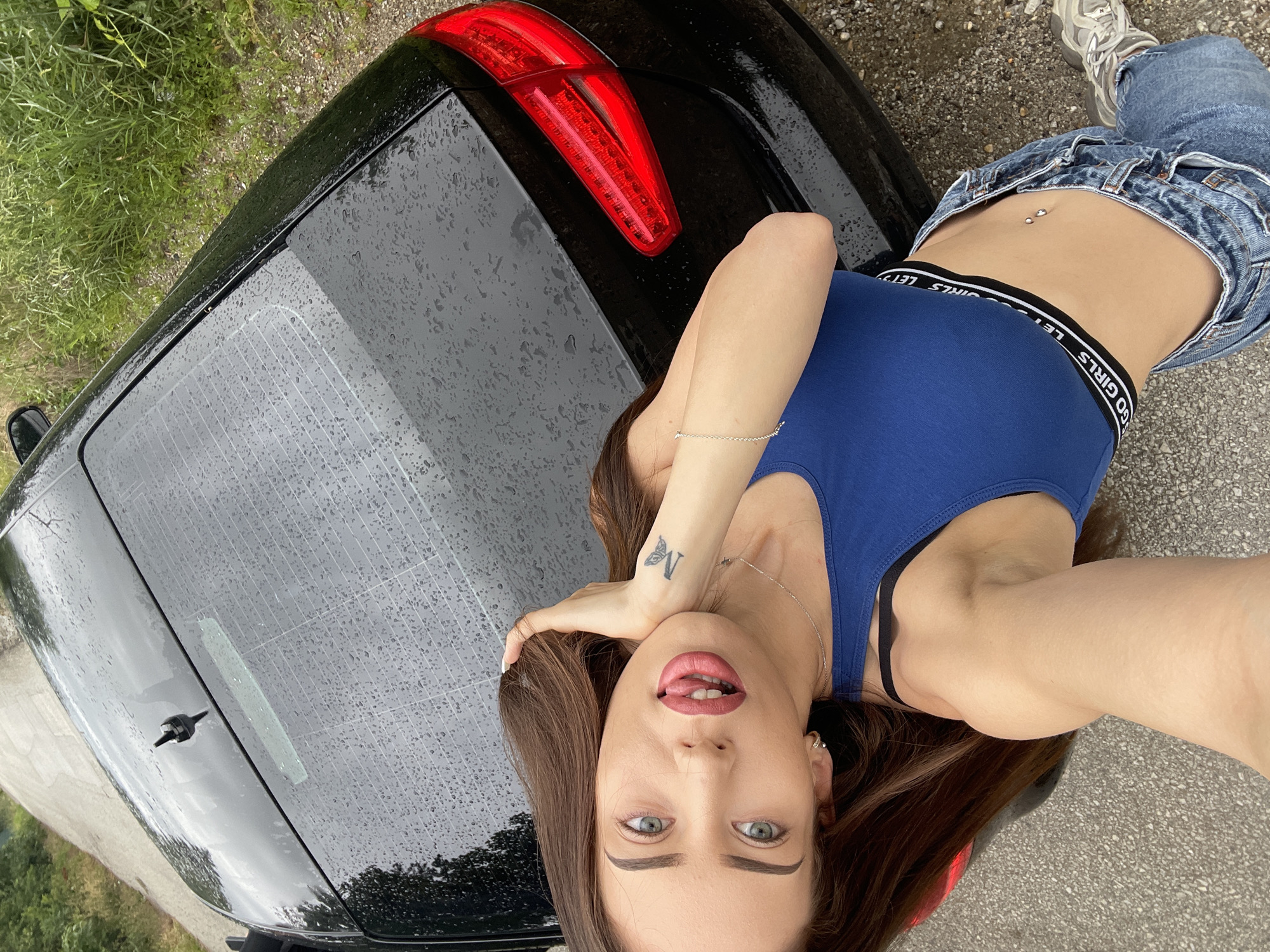 An overhead, high-angle medium shot captures a woman from the chest up as she leans against the rear of a black car. The car's rear windshield is wet with raindrops and appears to be a hatchback. A woman with long brown hair and light blue eyes is positioned in the lower left of the frame, her face tilted slightly upwards towards the camera. She is wearing a royal blue crop top with a black band that reads "LET'S GO GIRLS" and light blue denim jeans. Her right arm is bent, with her hand resting on her head, while her left arm is down her side. She has a tattoo on her left wrist of a butterfly with the letter "M" underneath. A silver necklace is visible around her neck, and she has a belly button piercing. The scene is outdoors, with a gravel road and lush green foliage in the background. The weather appears to be overcast or rainy, as indicated by the raindrops on the car. The location is identified as Unknown, Unknown.