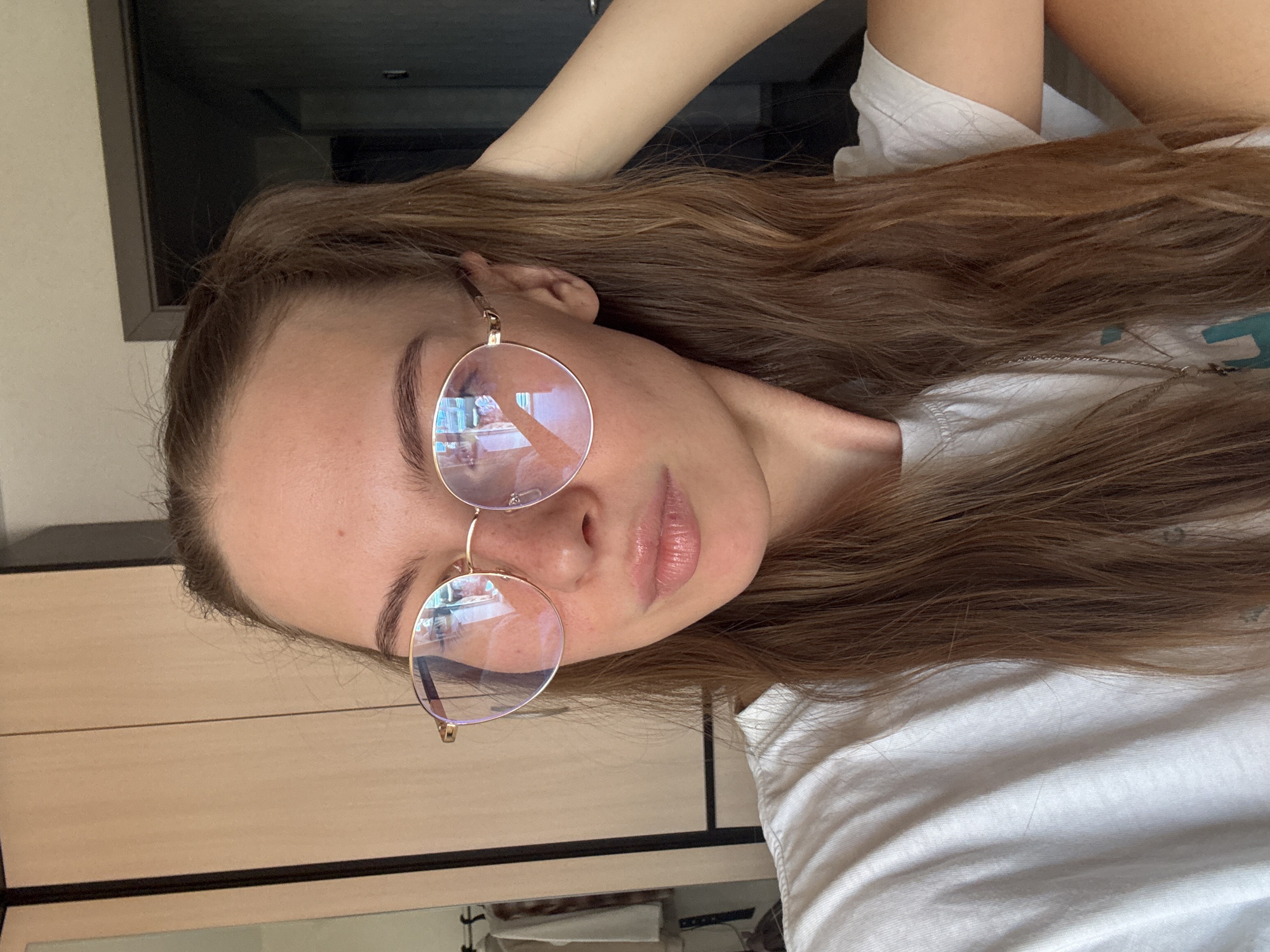 A young woman with long brown hair pulled into a ponytail, wearing round gold-rimmed glasses and a white t-shirt, is shown in a close-up, self-taken photograph. She is looking to her left, with her right arm behind her head. The background includes a closet with wooden doors and a glimpse of a doorway. The lighting suggests an indoor setting, possibly a bedroom. The image does not provide any specific information that would allow for determination of its location within the city of Perm, Russia.