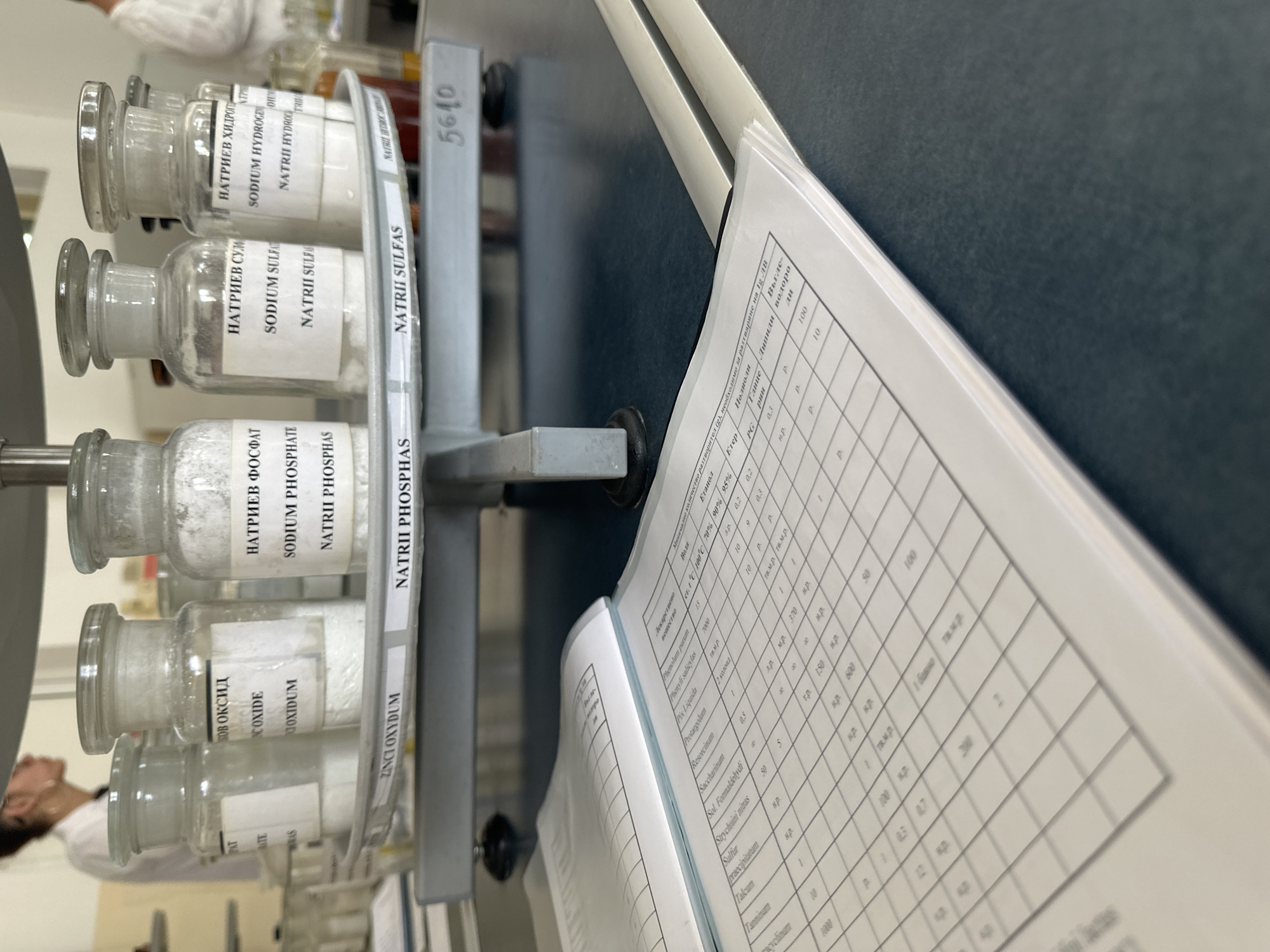 This media file captures an indoor scene in what appears to be a laboratory or chemistry classroom in Sofia, Bulgaria. The image is taken from a slightly angled perspective, looking across a dark tabletop.
In the foreground and midground, a set of clear glass reagent bottles, containing white powdery substances, are neatly arranged on a metallic rack. Each bottle features labels in both Cyrillic (Bulgarian) and Latin script, identifying their contents. Visible labels include "НАТРИЕВ ХИДРОГЕН / SODIUM HYDROGEN / NATRII HYDROC...", "НАТРИЕВ СУЛЬФАС / SODIUM SULFAT / NATRII SULFAS" (Sodium Sulfate), "НАТРИЕВ ФОСФАТ / SODIUM PHOSPHATE / NATRII PHOSPHAS" (Sodium Phosphate), and "ЦИНКОВ ОКСИД / ZINC OXIDE / ZINCI OXYDUM" (Zinc Oxide). The metallic rack itself bears the numerical label "5610".
To the right, a white printed document or textbook is open on the table, displaying a grid or table with chemical names and various numerical data, likely related to solubility or properties of substances. Column headers visible on the document include "Лекарствено вещество" (Medicinal substance), "Вода" (Water), "Етанол" (Ethanol), "Етер" (Ether), "Глицерин" (Glycerin), "Липиди" (Lipids), and "Въглеводороди" (Hydrocarbons), along with percentages like "70%" and "90%". Specific chemical names such as Phenolum purum, Phenyli salicylas, Protargolum, Resorcinum, Saccharinum, Strychnini nitras, Sulfur praecipitatum, Talcum, and Tanninum are clearly legible within the table.
In the background, partially out of focus, a person wearing a white lab coat is visible, suggesting an active presence in the lab environment. The overall impression is one of a scientific or educational setting, possibly during a practical lesson or experiment. The lighting is artificial, typical of an indoor laboratory setting.