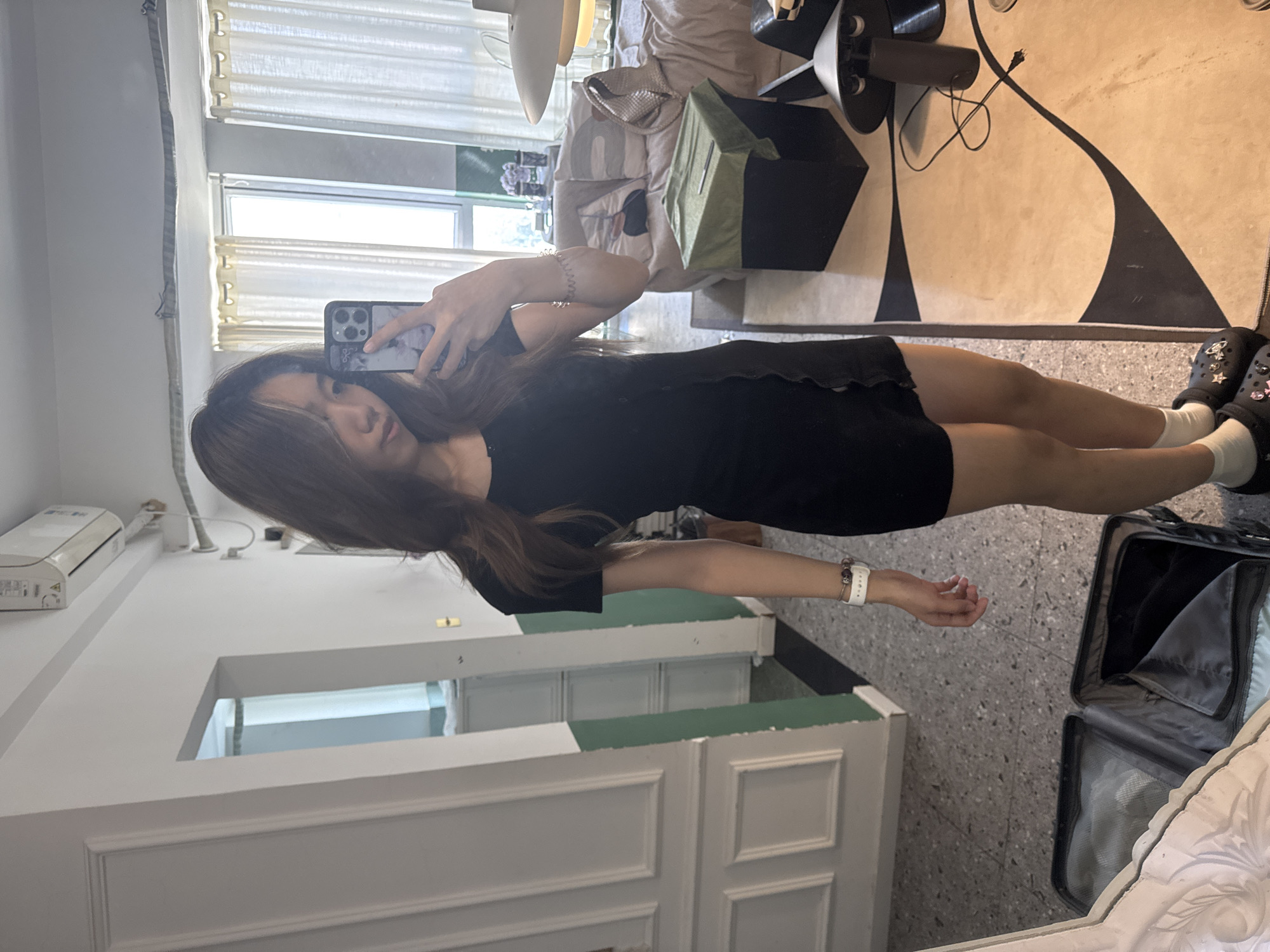 A young woman is taking a mirror selfie in what appears to be an apartment in Liangjiang, China. She is wearing a black dress, white socks, and embellished black shoes. She is holding a smartphone to take the picture. In the background, there is an open suitcase on the floor, a white couch with some items on it, a black side table with a lamp, and a large rug with a black and white pattern. A white vanity with green trim is also visible, as well as curtains over a window.
