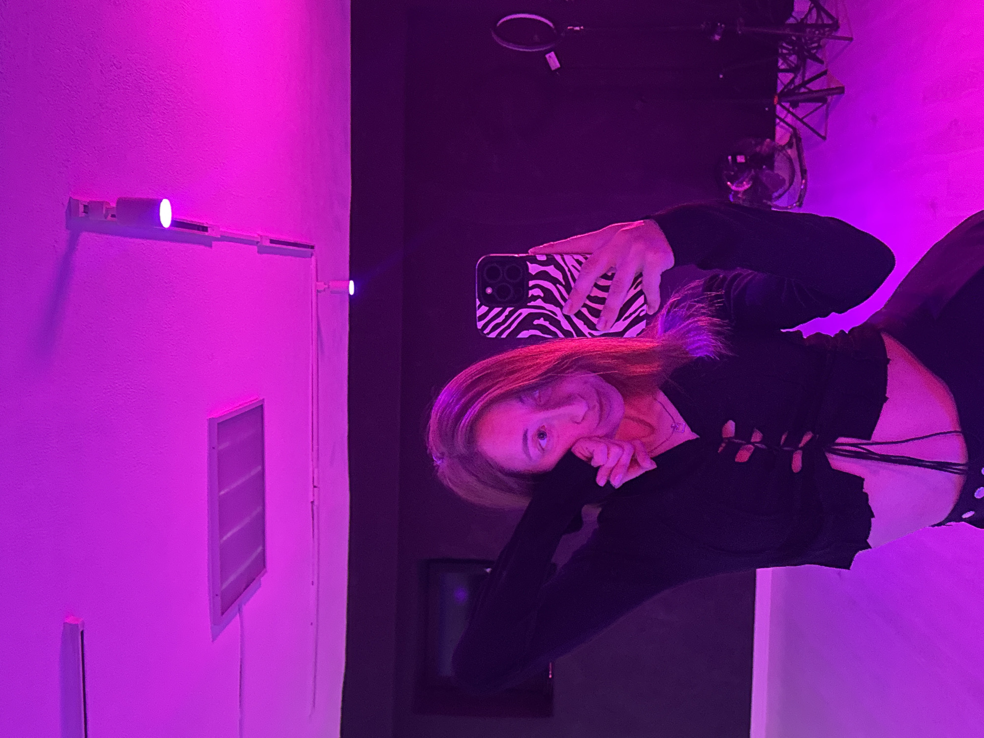 The image captures a female-presenting individual taking a selfie, likely in a mirror or reflective surface, within an indoor setting illuminated by vibrant magenta and purple lighting.
**Subjects:** The main subject is the person, whose long, light brown hair is cast in purple by the ambient light. She is wearing a dark, possibly black, long-sleeved top with lacing or cut-out details at the midriff. Her right hand partially covers her face, revealing one eye with a neutral or slightly playful expression. She holds a smartphone with a distinctive black and white zebra-print case.
**Objects:** The smartphone held by the subject features multiple camera lenses. On the textured wall behind her, several white, linear LED light fixtures are mounted, with one emitting a particularly bright purple glow. A rectangular, louvered vent is also visible on this wall. In the darker background area, indistinct equipment can be seen, including what appears to be a microphone stand, a small fan or air circulator mounted near the ceiling, and a dark rectangular monitor or screen.
**Scene and Setting:** The scene is an indoor room, possibly a studio or a dedicated creative space, characterized by a predominantly magenta and purple artificial light source. One wall is brightly lit in this hue, while other areas recede into darker shades of purple and black. The wall surface has a visible texture. The lighting creates a stylized and moody atmosphere.
**Activity or Event:** The person is engaged in the act of taking a selfie, with her attention directed towards the phone she is holding, which is pointed back at her.
**Notable Details:** The most striking detail is the pervasive magenta/purple lighting, which gives the entire scene a dramatic and artificial feel, making the time of day indeterminate. The presence of studio-like equipment in the background suggests the location might be a recording studio, a photography setup, or a content creation space. No discernible text is visible in the image. The location context is Chaikovsky, Russia.
