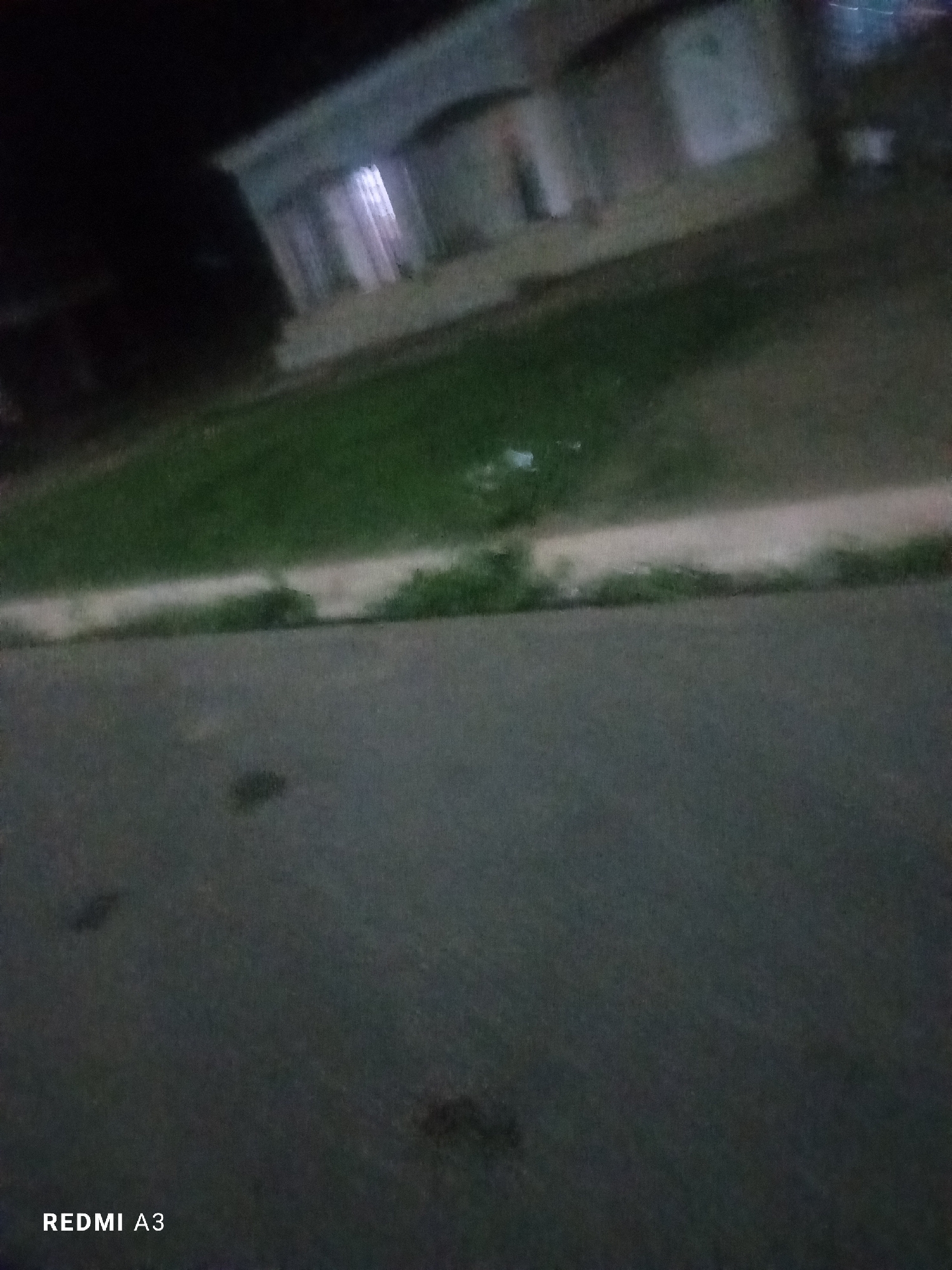 The image shows a blurry nighttime view of a street in Jalingo, Nigeria, with a building in the background. The building appears to be a residential or commercial structure with illuminated windows. A grassy area is visible between the street and the building, separated by a lighter colored strip. The street itself is dark asphalt with some faint marks. The bottom left corner of the image displays the text "REDMI A3".