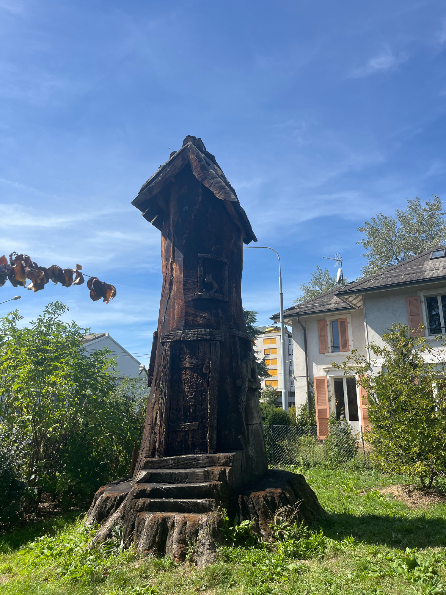 The image shows a large, elaborately carved wooden structure resembling a miniature house or tower, possibly a sculpture, made from a tree stump. The structure is dark brown and weathered, with a pitched roof. Steps are carved into the base of the stump, leading up to a small opening or window on the front of the structure. In the background, there are green trees, a residential building with windows and shutters, and a clear blue sky with some wispy clouds. A streetlamp is visible to the left of the wooden structure. The scene appears to be an outdoor setting in a residential area. The image was taken on a sunny day.