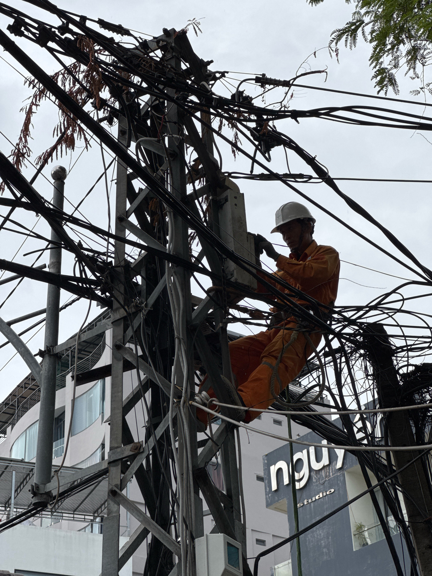 A male worker in an orange jumpsuit and white hard hat is seen perched on a complex network of electrical wires and poles, in what appears to be a densely populated urban area of Nha Trang, Vietnam. He is actively engaged in work on a junction box, using tools. The surrounding environment is characterized by a heavy tangle of black and grey electrical cables, some adorned with dry, brown foliage, and bare, uninsulated wires. In the background, a modern, multi-story building with curved windows and a signage reading "nguy studio" is visible, suggesting the location is in a commercial or residential district. The sky is overcast, contributing to a muted lighting condition.