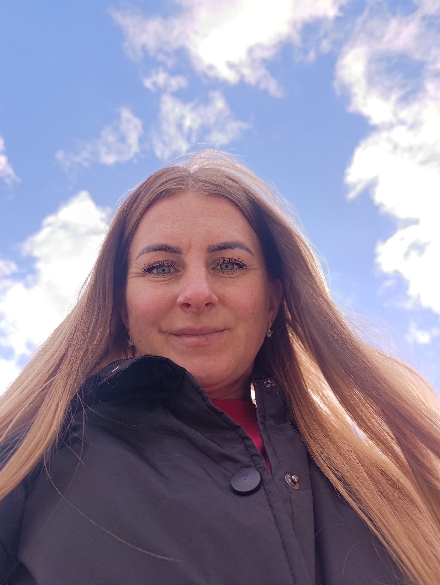 A woman with blue eyes and long, reddish-blonde hair is centered in the frame, looking down at the camera. She is wearing a black jacket with a visible button and snap fasteners, and a glimpse of a red top can be seen beneath the jacket. The background is a bright blue sky filled with puffy white clouds. The location is Unknown, Unknown.