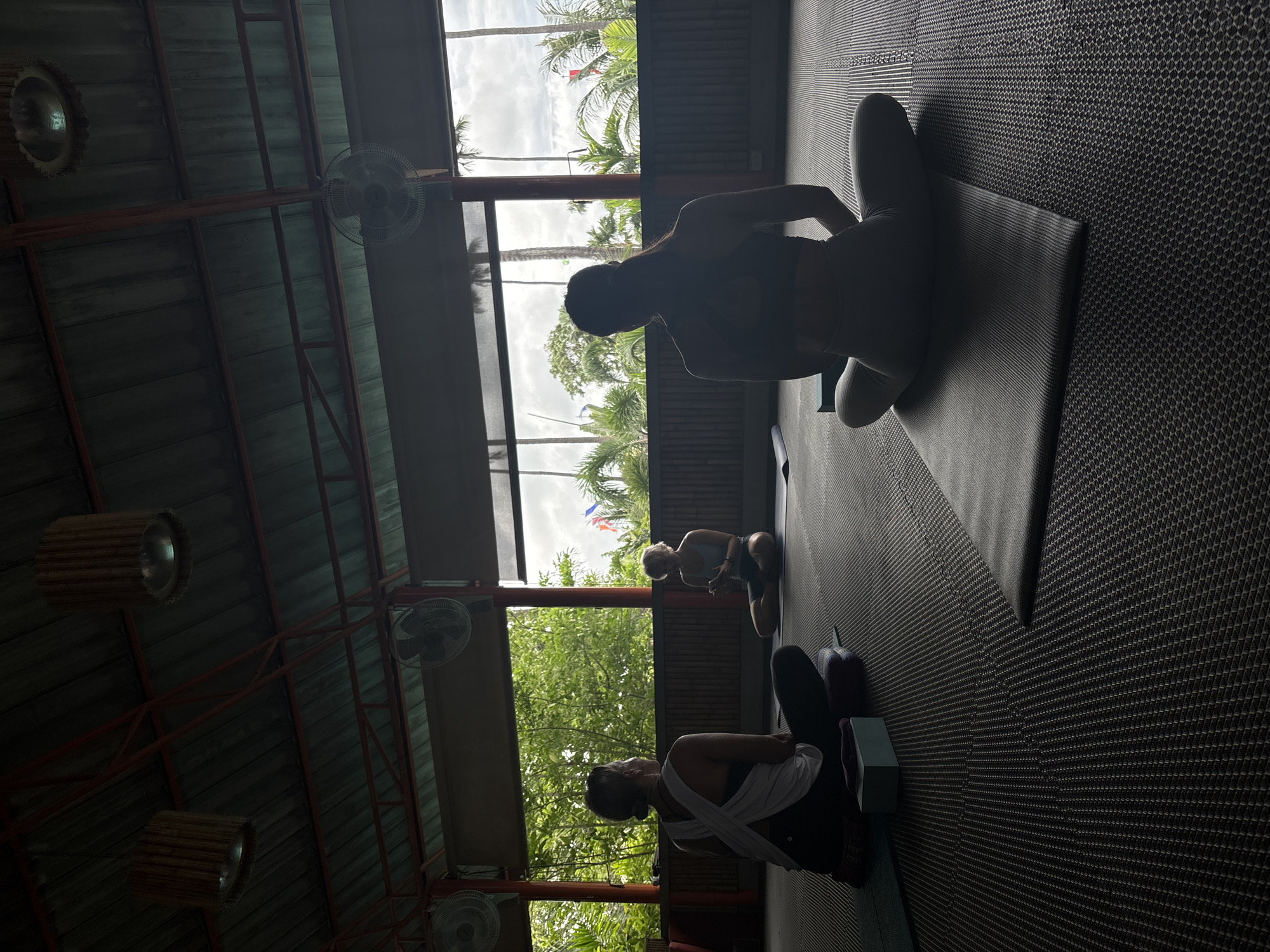 Captured in what appears to be an indoor yoga or exercise studio in Rawai, Thailand, the image showcases three individuals engaged in seated poses on yoga mats. The setting is an open-air structure with a roof supported by exposed red beams, fans hanging from the ceiling, and woven lamps. Daylight filters in through the open walls, revealing glimpses of lush greenery outside, characteristic of Thailand’s tropical environment. The people are positioned linearly in the frame. One has a white shirt on and the other two are in dark clothes. They appear to be participating in a yoga or meditation session.
