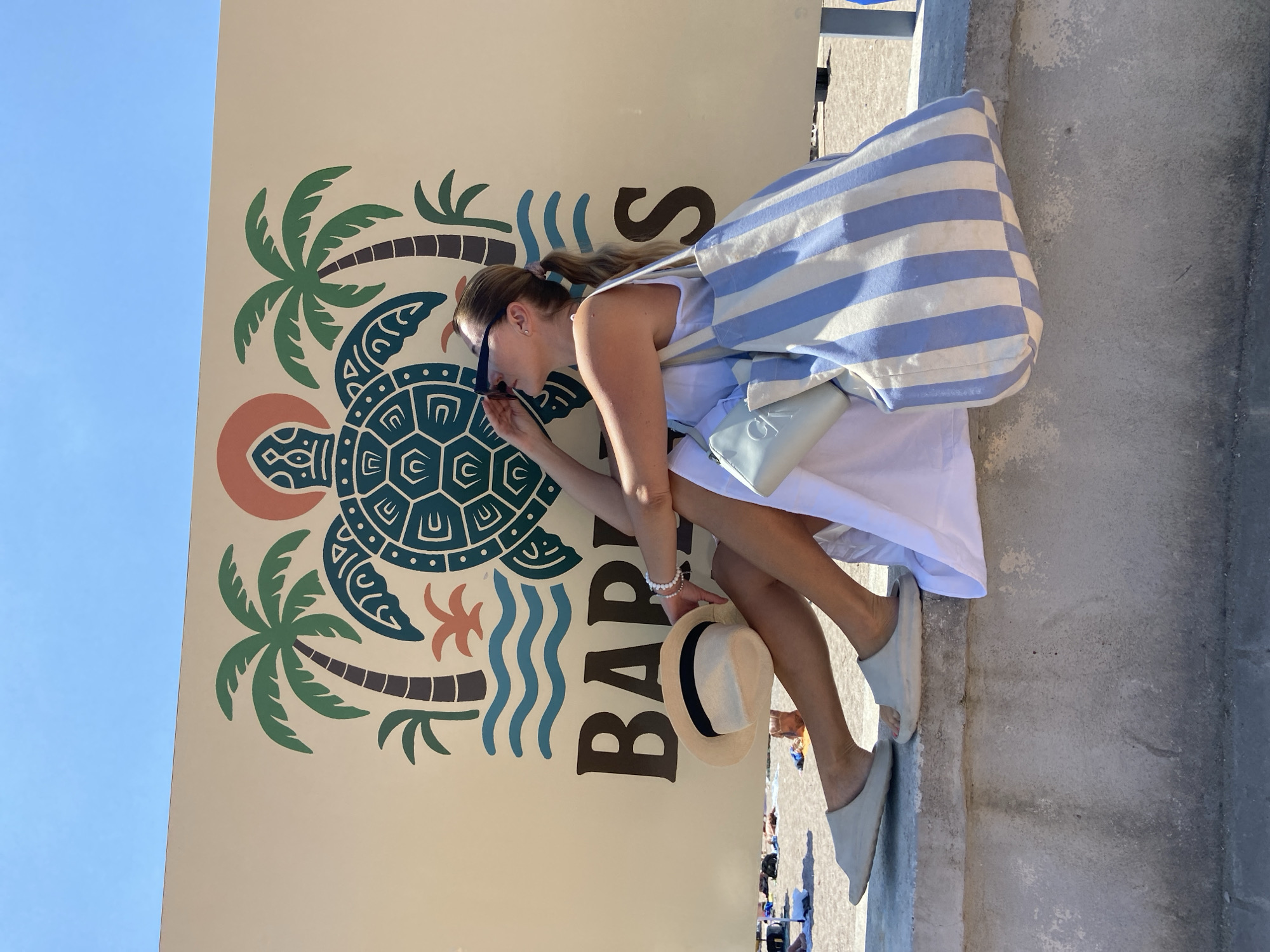In Prijevor, Montenegro, a woman sits on a concrete ledge in front of a large, beige banner with a stylized graphic of a turtle, palm trees, and waves. Text on the banner reads "BARS". The woman is wearing a white dress, sunglasses, and light-colored slip-on sandals. She is holding a straw hat with a black band in her left hand and has a light blue clutch and a large blue and white striped tote bag beside her. Her hair is pulled back in a ponytail. The background shows a beach scene with people and umbrellas. The sky is clear and blue.