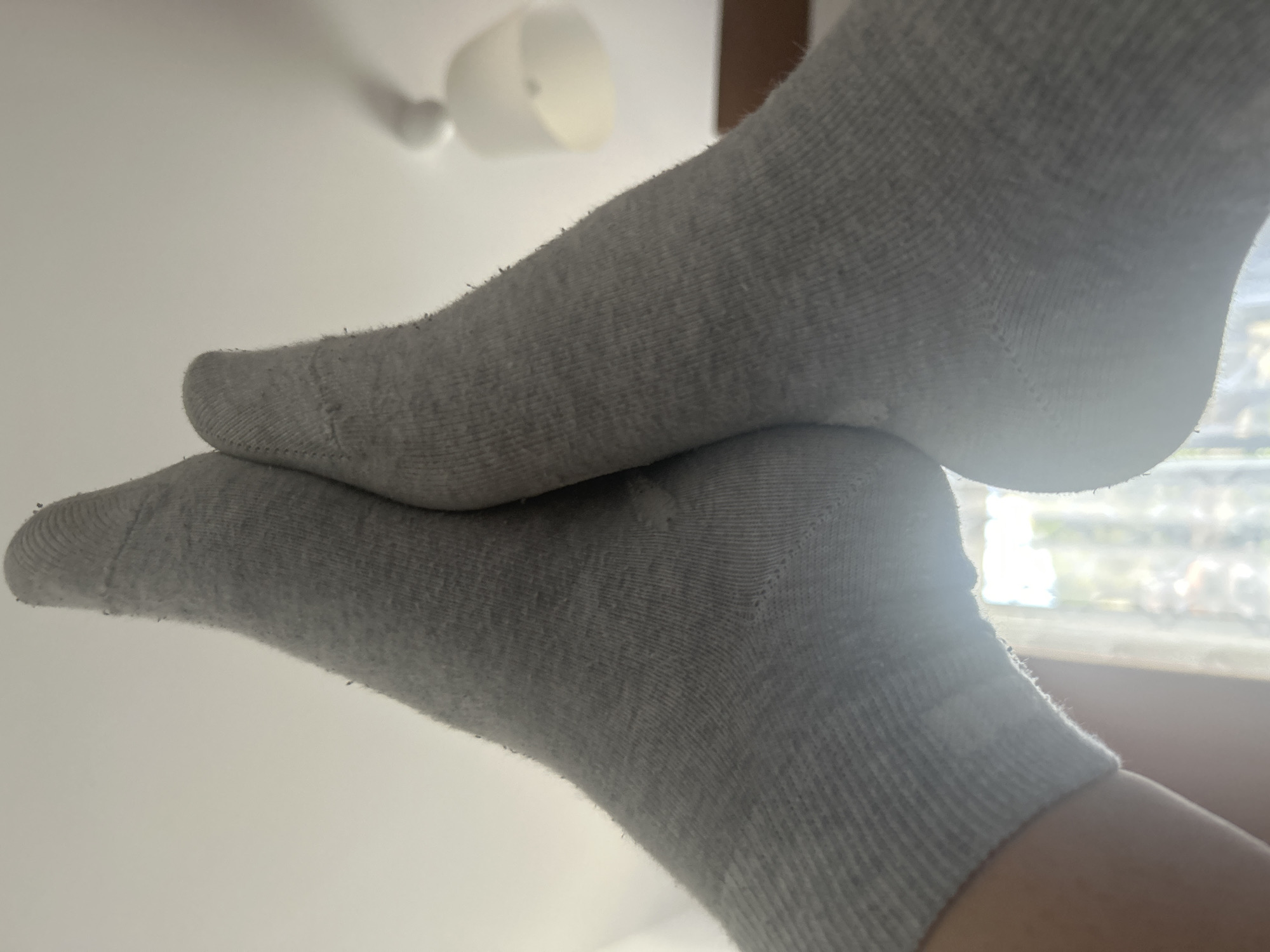 The image shows two feet wearing gray socks resting on each other. The background is a white wall with a light fixture and a window with a blurry outdoor view. The location cannot be determined from the image.