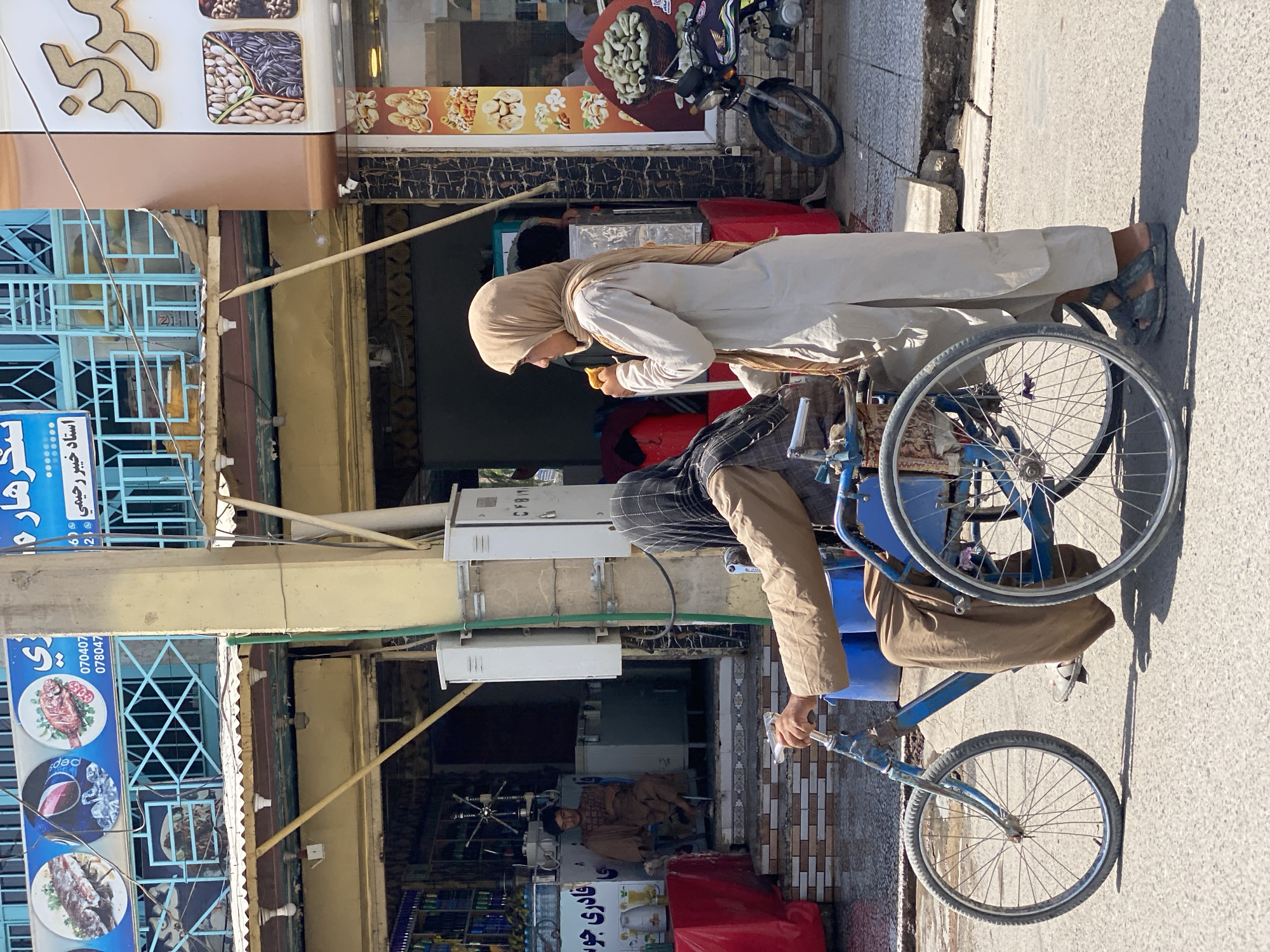 This is an eye-level, medium shot of a street scene in Kandahar, Afghanistan. In the foreground, a person in a wheelchair, wearing a brown tunic and headscarf, is being pushed by another person wearing a tan tunic and light brown headscarf. The person in the wheelchair is holding the handles of the wheelchair. The person pushing is holding a piece of food. The wheelchair is a blue, three-wheeled vehicle with a patterned seat. The environment is a paved street with shops and signage in the background. One shop has a blue sign with images of fish and a beverage. Another shop has a tan and brown sign with images of nuts and dried fruit. A motorcycle is parked to the right of the frame. The lighting suggests it is a sunny day.