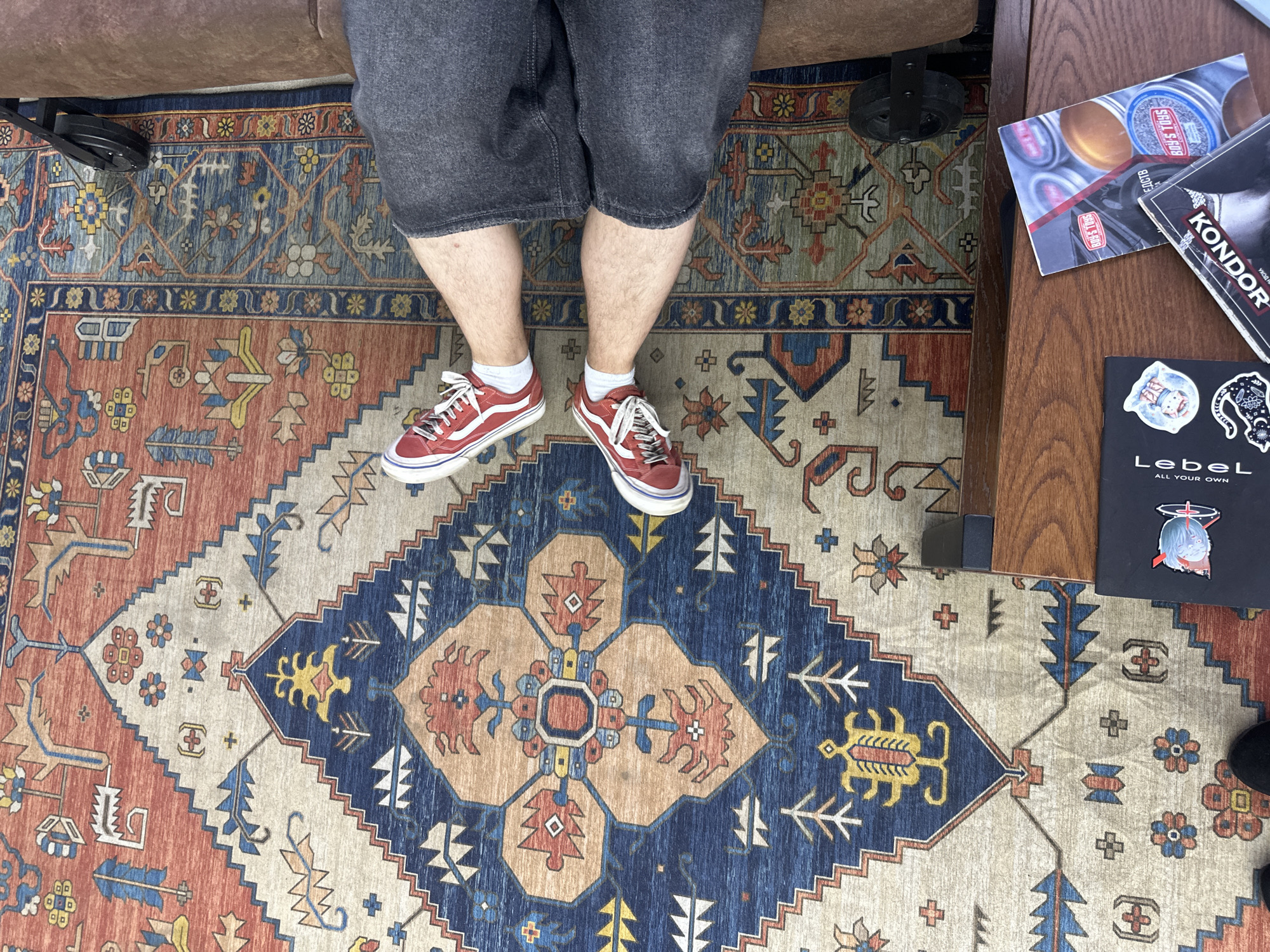 This media file captures a low-angle, top-down shot of a person's legs and feet adorned with red Vans sneakers, standing on a richly patterned oriental rug. The person is wearing dark denim shorts and white socks. The rug displays intricate geometric designs in shades of red, blue, beige, and yellow. To the right of the rug, a portion of a wooden table is visible, holding several items, including what appears to be a magazine or booklet with the text "BOY'S TOYS" and "KONDOR" on it. A smaller black booklet with the brand name "LebeL" and "ALL YOUR OWN" is also present, featuring stickers of cartoon characters. In the background, a brown leather couch and a black chair with a wheel are partially visible. The scene suggests an indoor setting, possibly a lounge or a store, with a casual and relaxed atmosphere. The lighting indicates it could be daytime, with no specific weather cues. The presence of the products on the table might suggest a retail or promotional context.