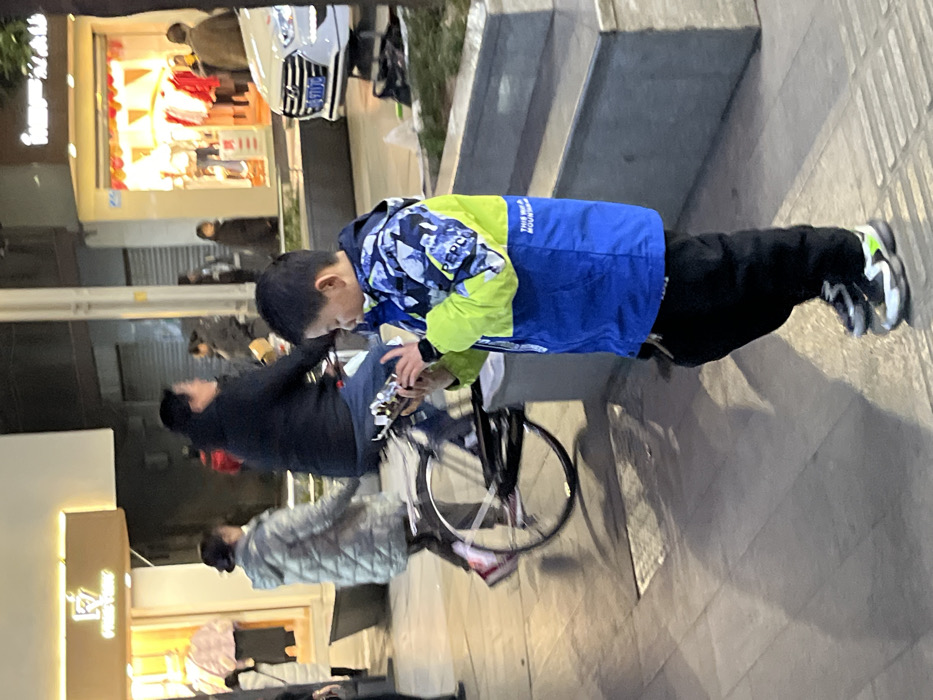 An East Asian male child stands on a tiled sidewalk in Jiangjin, China, intently viewing a horizontally held smartphone. He wears a blue, neon green, and camouflage-patterned jacket with "PEPPA" text on the sleeve and "THIS IS A MOUNTAIN" on the front, black trousers, and black, white, and neon green sneakers. A dark band is visible on his right wrist. In the midground, an East Asian female cycles a bicycle from left to right, wearing a dark jacket. To her left, another East Asian female walks away from the camera, wearing a grey puffer jacket and dark trousers. In the background, brightly lit storefronts are visible, including one with a "FRIDE FEIRY" sign displaying merchandise and another with red decorations and clothing items. A white sedan is parked to the right, partially visible. The scene is illuminated by artificial light sources, indicating an evening or nighttime setting in an urban commercial area.