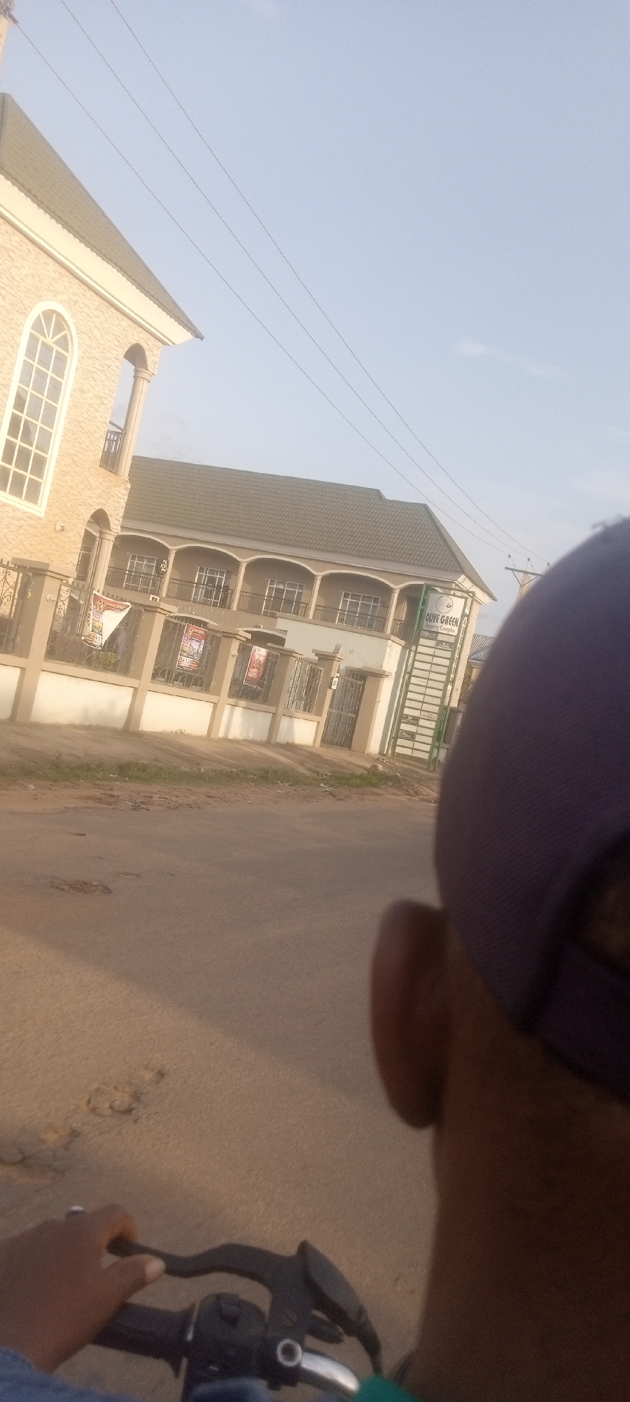 The image shows a street scene in Narayi, Nigeria. In the foreground is a person riding a motorcycle, with a partial view of their hand on the handlebar, part of their blue shirt, and the back of their head with a blue cap. The person is traveling on a paved road with some potholes and patches.
In the background, there are two buildings separated by a fence. The building on the left has an arched window and a light-colored exterior. The building on the right has a green roof, a second-story balcony, and is labeled "OLIVE GREEN Shopping Complex." Utility poles and wires are visible above the buildings. Banners are affixed to the fence.