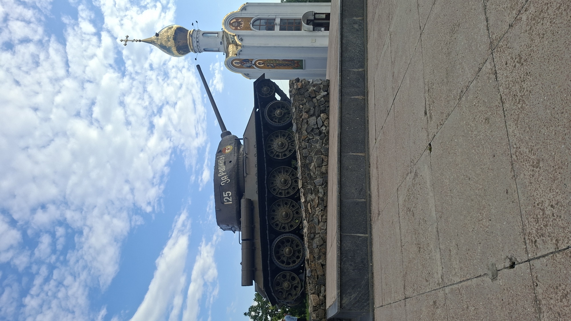 A T-34 tank monument, painted dark green, stands on a stone embankment with a church to its right. The tank has the number "125" and the Cyrillic inscription "ЗА РОДИНУ!" (For the Motherland!) painted on its side, along with a red star. The church features a golden dome topped with a cross and decorative mosaics on its facade. The scene is set against a bright blue sky with scattered white clouds. In the foreground, a concrete paved area with visible cracks and some small weeds is visible. A person in a light-colored hat is partially visible on the left side of the image, near some greenery. The monument is located in Tiraspol, Moldova.