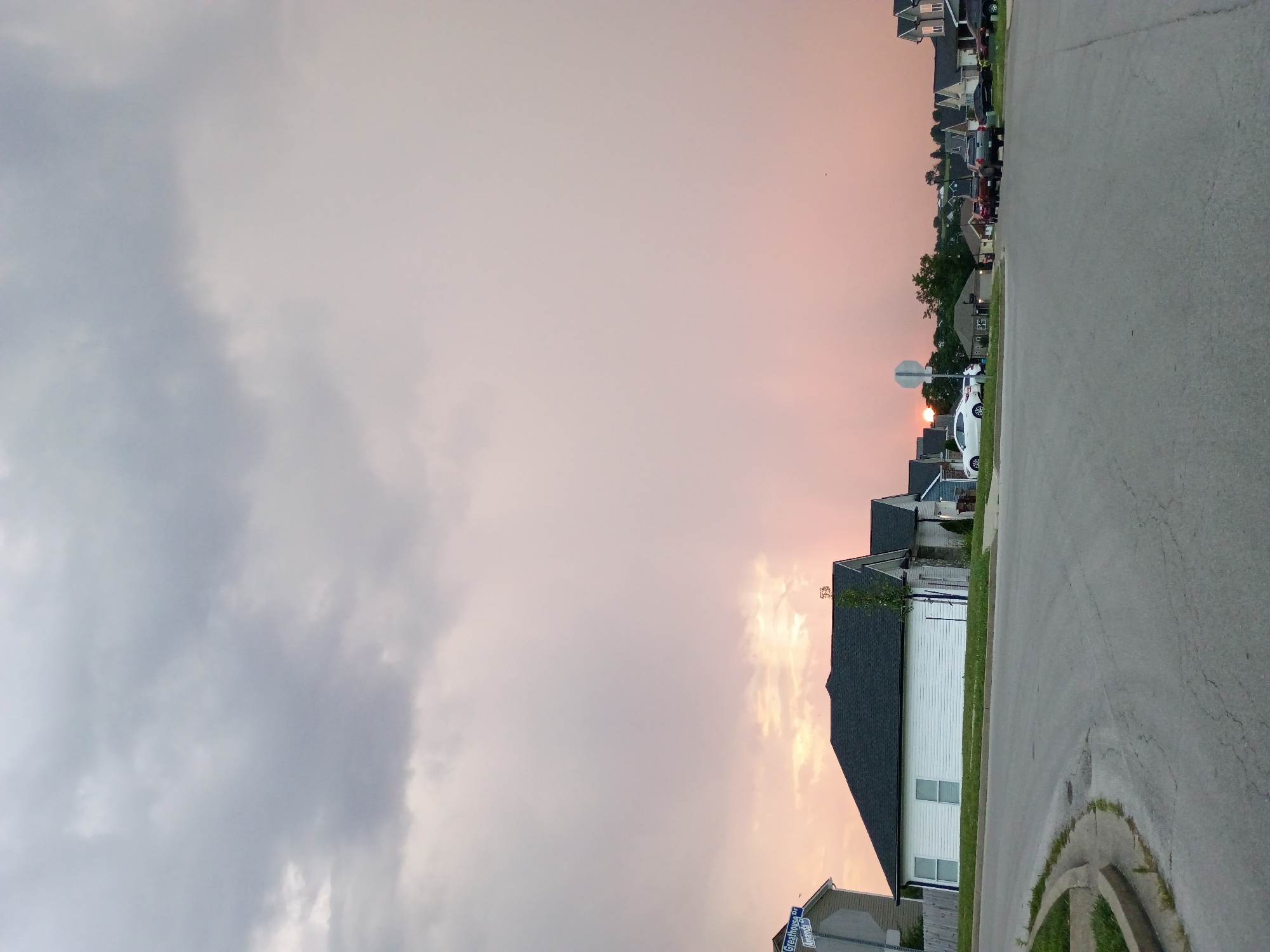 A street sign reads "Greathouse Dr" and "Amanda St" at the intersection of two paved roads. In the background, the sun is setting behind a line of houses, casting a pinkish glow on the clouds. The sky is overcast with heavy gray clouds. A white, single-story house with a dark roof is situated on the left side of the image, with two windows visible. Several cars are parked in front of houses in the distance. The scene appears to be a residential neighborhood.