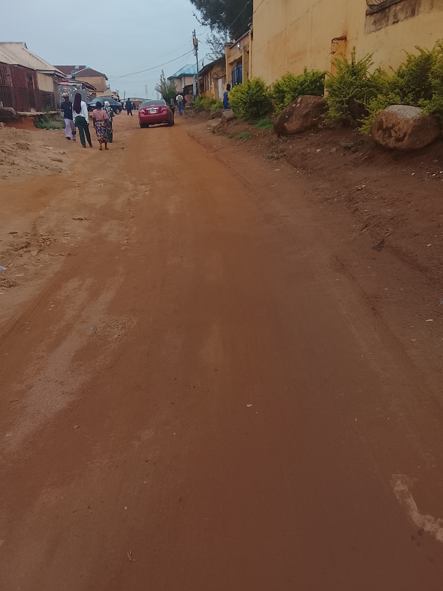 The image depicts an unpaved, red dirt road in what appears to be a residential area. A group of people are walking away from the camera on the left side of the road, with a red car parked further ahead. Buildings line both sides of the road, some constructed of brick and others painted yellow. Green bushes and rocks are positioned along the right side of the road near the buildings. The sky is overcast. The location appears to be a neighborhood in Jos, Nigeria, characterized by its unpaved roads and modest buildings. The scene is likely a common occurrence within the city.