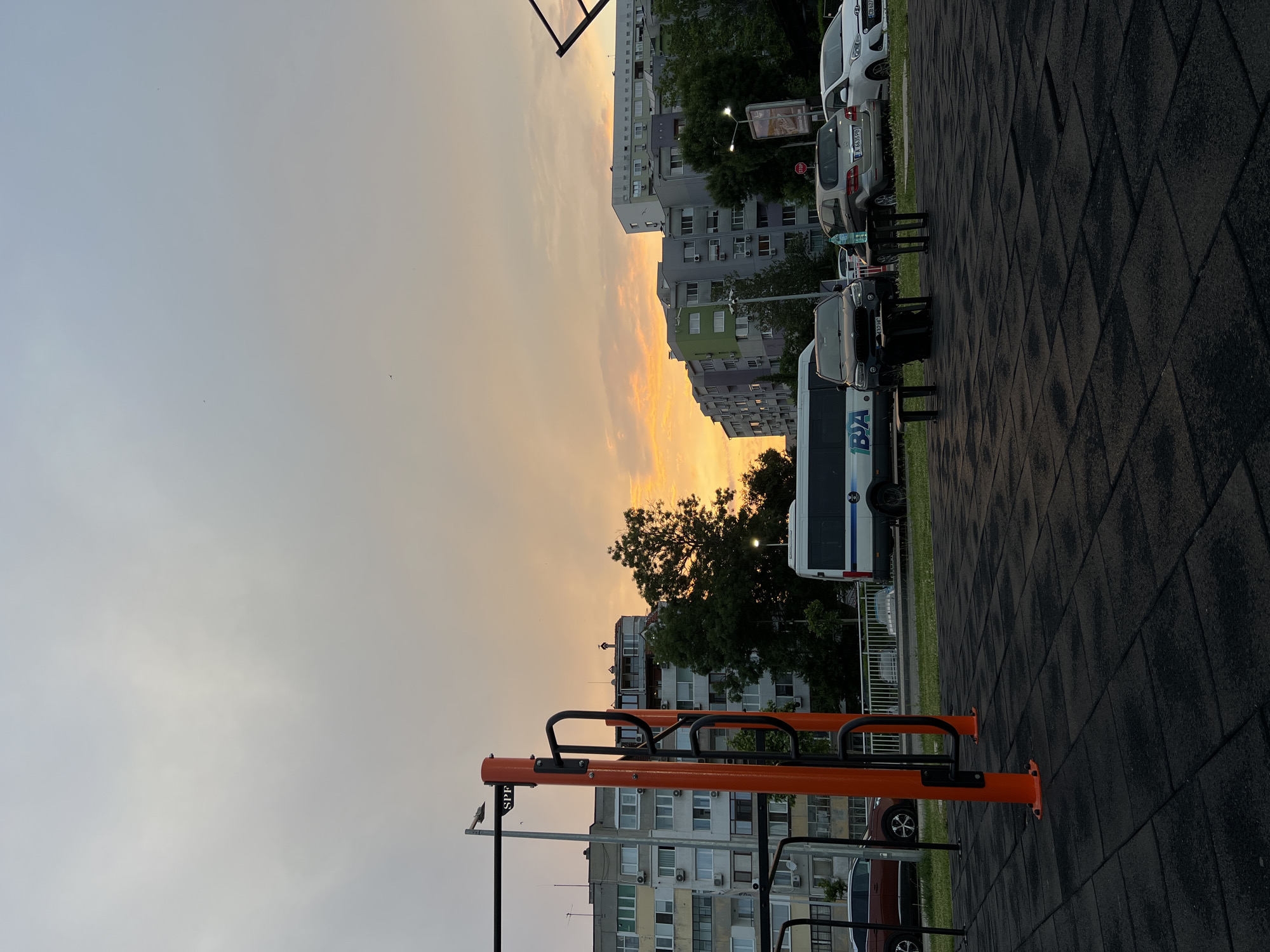 The image captures an urban landscape at either sunrise or sunset, characterized by a warm, gradient sky. The time of day is evident from the vibrant orange and yellow hues dominating the right side of the sky, gradually fading into lighter grey and blue tones towards the left. The light casts a soft glow on the upper parts of distant buildings and clouds, while the foreground and midground elements appear somewhat shadowed.
In the foreground, a modern outdoor exercise area is visible, featuring orange and black metal calisthenics equipment. The ground beneath the equipment is covered with dark, square rubber tiles. Part of the equipment shows the partial text "SPF".
Beyond the exercise area, a street or parking lot stretches across the midground. Several vehicles are parked along it, including a white van with a partially visible license plate "CA 6570 HT", a silver car displaying the license plate "B 6335 PH", and a darker car with "H 1247 HP" on its plate. A white and blue bus, marked with the text "BPA", is also present. A red "STOP" sign is visible on a pole behind the silver car. Lush green trees are interspersed among the vehicles and buildings.
In the background, multi-story residential buildings with numerous windows and balconies line the street. Some buildings have light-colored facades, while others show hints of green and grey. Small birds are faintly visible flying against the vast sky. No people are seen in the frame. The overall scene suggests a quiet moment in a residential area of Burgas, Bulgaria, during a transitional light period of the day.