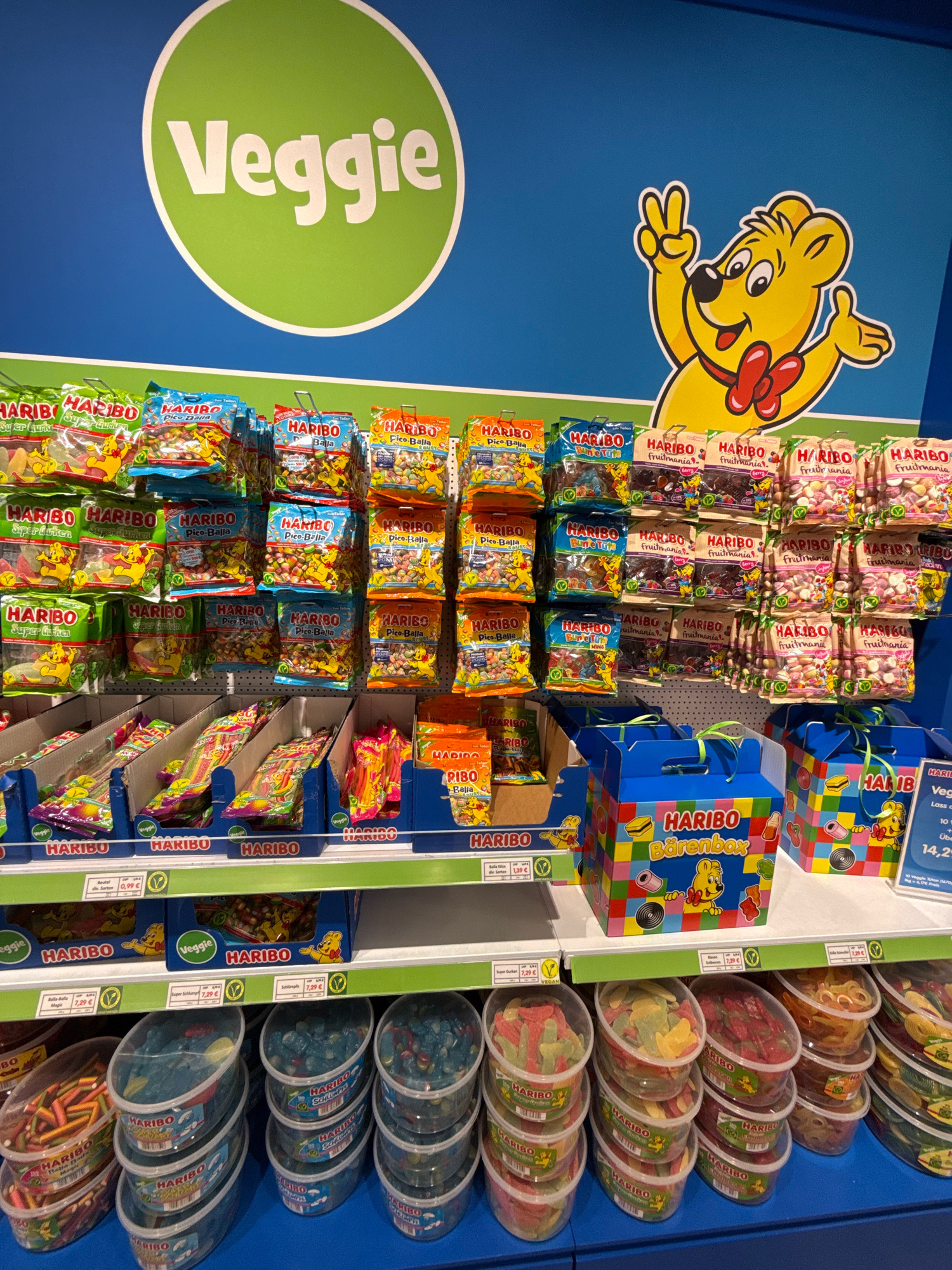 The image shows a display of Haribo brand "Veggie" gummy candies in a store. The display is organized on shelves and features numerous bags and tubs of candy, predominantly featuring the Haribo bear mascot and green leaf symbols indicating vegetarian options. In the background, a large green circle with the word "Veggie" in white lettering is visible on a blue wall, along with a larger illustration of the Haribo bear giving a peace sign. On the right side of the image, a price tag for "Veggie" candies is partially visible, listing prices in Euros. The overall scene suggests a retail environment, likely a confectionery aisle in a supermarket or candy store. Based on the presence of the "AutoMuseum Volkswagen" sign visible in the distance through the store window, this scene is located in Wolfsburg, Germany, within the Volkswagen Autostadt complex. No people are visible in the image.