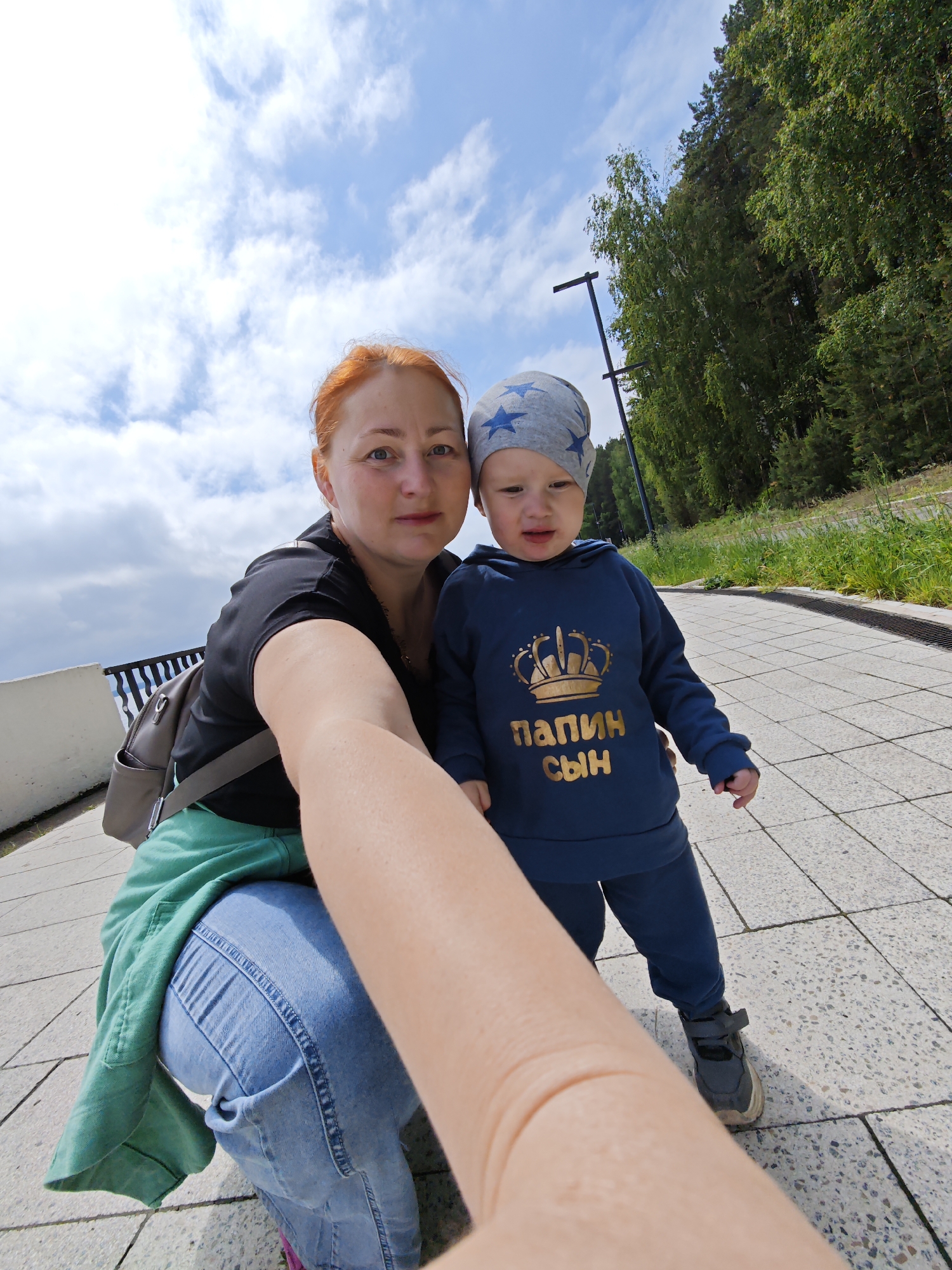 An outdoor selfie taken in Chaikovsky, Russia, features a woman and a young child, likely a mother and her son, on a sunny day. The woman, with red hair, is seen crouching, wearing a black t-shirt, light blue jeans, and a green jacket tied around her waist. A gray backpack is visible on her shoulder. She extends her right arm to capture the photo. Standing beside her is a young boy, a toddler, dressed in a gray beanie adorned with blue stars, a navy blue hooded sweatshirt, matching sweatpants, and gray and white sneakers. The sweatshirt prominently displays the Russian text "ПАПИН СЫН" (Dad's Son) in gold lettering.
The setting is a paved pathway, possibly a promenade or park path, with a dark metal railing visible to the left. The background is dominated by a lush, dense forest of tall green trees under a bright blue sky dotted with scattered white clouds. The clear, bright light indicates a daytime setting with pleasant weather. Both subjects have neutral to slightly smiling expressions, suggesting a relaxed, casual outing.