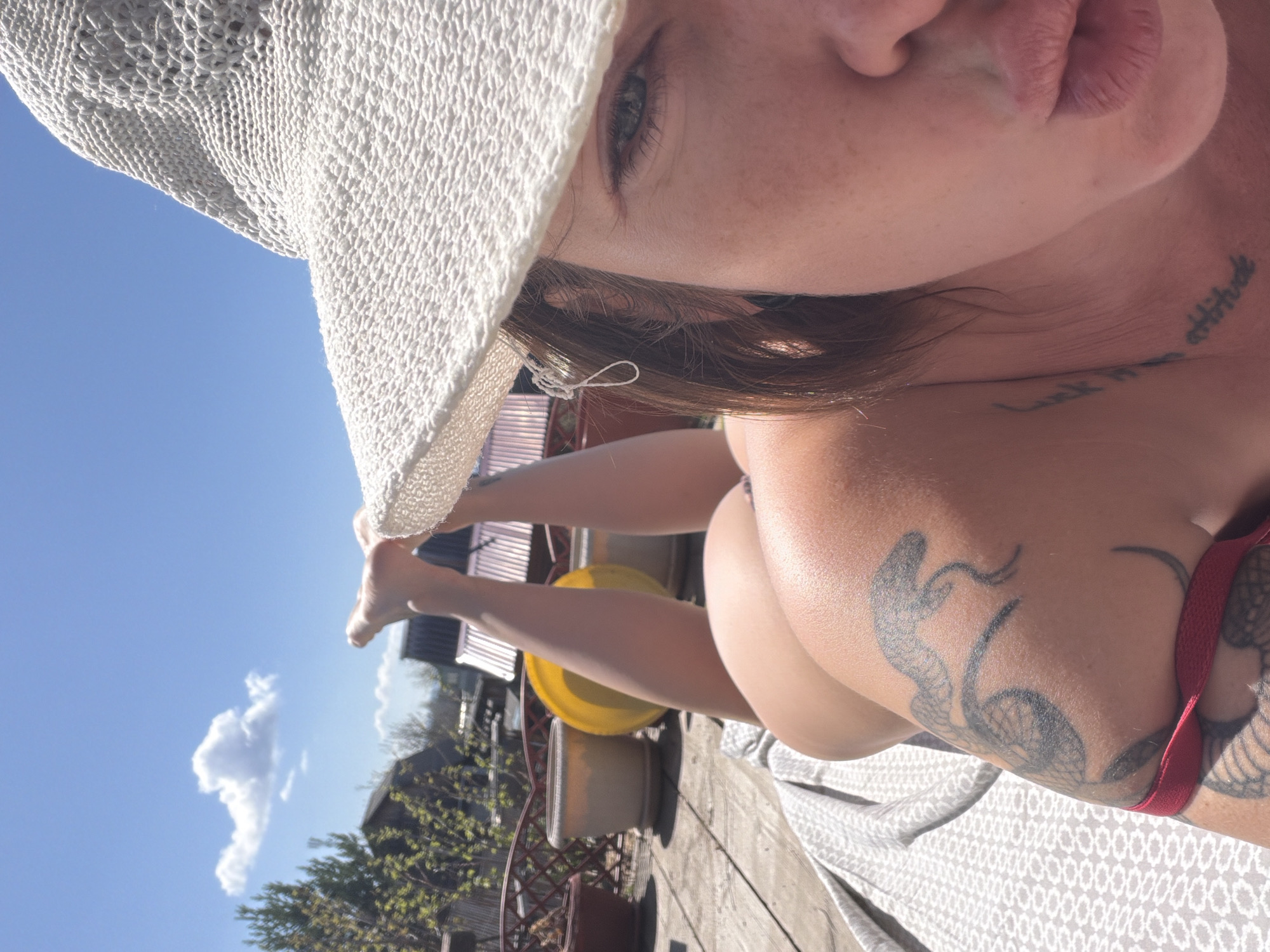 A photograph taken outdoors in Zheleznodorozhny, Russia, depicts a light-skinned individual with brown hair reclining on a patterned white surface, possibly a blanket or sheet. The subject is wearing a white crocheted hat that partially obscures their face. The subject has a tattoo on their neck, a snake tattoo on the upper arm. There is a red strap visible, possibly part of swimwear. In the background, there's a wooden deck with some potted plants, a yellow object, and a partial view of buildings, trees, and a blue sky with a cloud. The photo is angled and the subject's face is close to the camera.