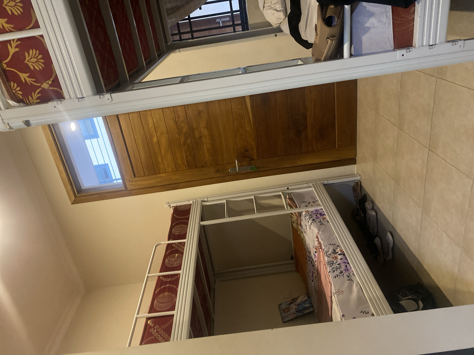 A view from the top bunk of a bunk bed in a room in Dar es-Salaam, Tanzania, looking at a door, a window, and the bottom bunk of the bunk bed. There is bedding on the bottom bunk, a picture on the wall next to it, and shoes and a bag near the bottom bunk. The city of Dar es-Salaam is presumed based on the style and furnishings of the room being consistent with images of accommodations in Dar es-Salaam.