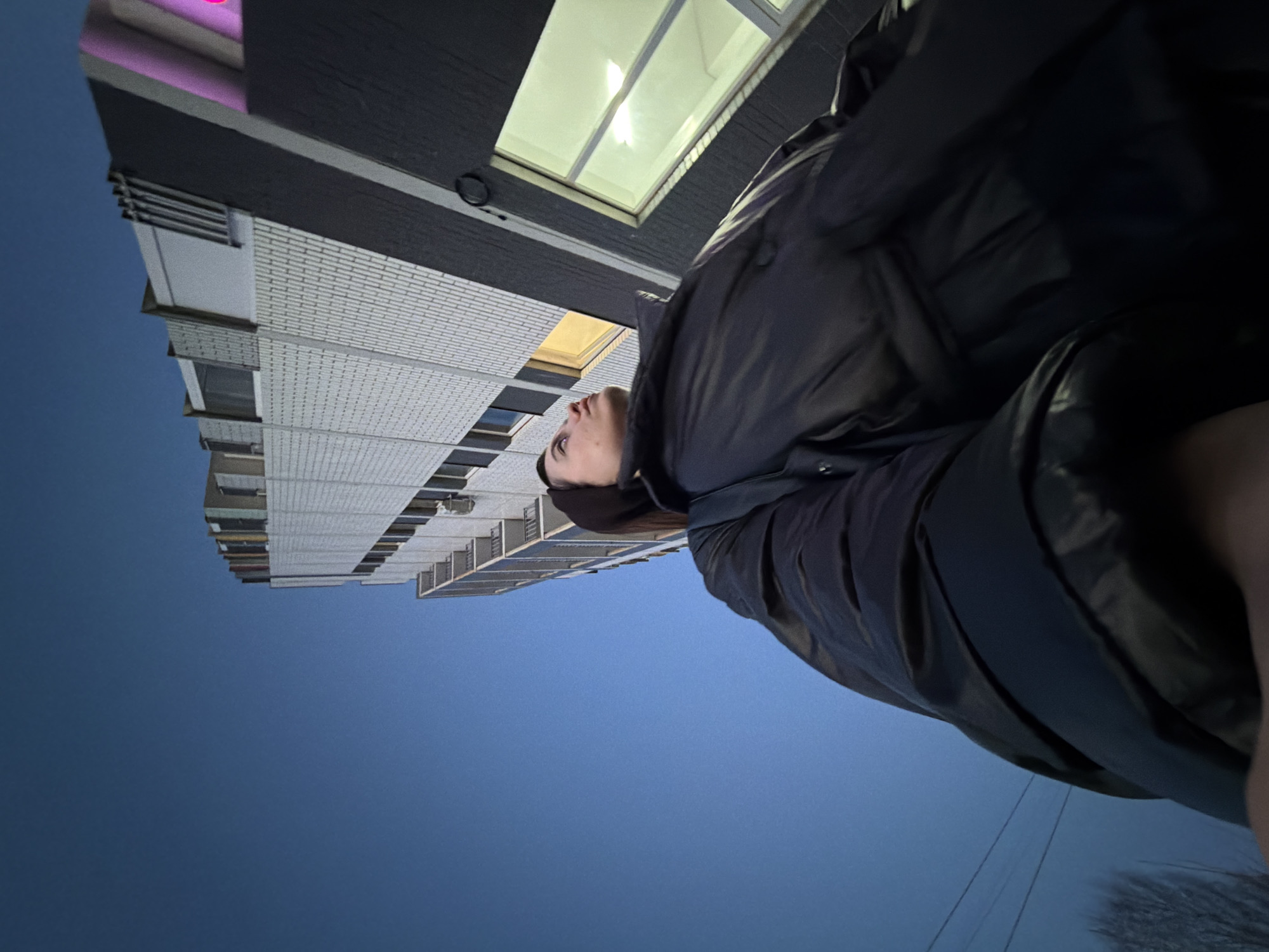 A person is standing in Chaikovsky, Russia looking up at a tall, white brick building with dark grey accents, which features several rows of windows. The time of day appears to be dusk or early evening, as the sky is a dark blue and lights are on inside the building. The person is wearing a dark-colored jacket and appears to be the one taking the photograph from a low angle. Bare trees and power lines are visible in the distant background, partially obscured by the person's jacket.